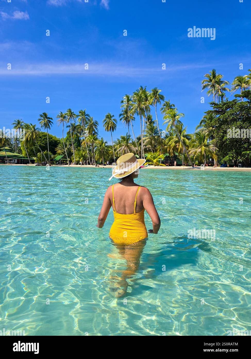 Una donna con un costume da bagno giallo passeggia attraverso le tranquille acque turchesi di Koh Kood. Circondata da palme ondeggianti e dal sole luminoso, gode di un momento sereno in paradiso. Foto Stock