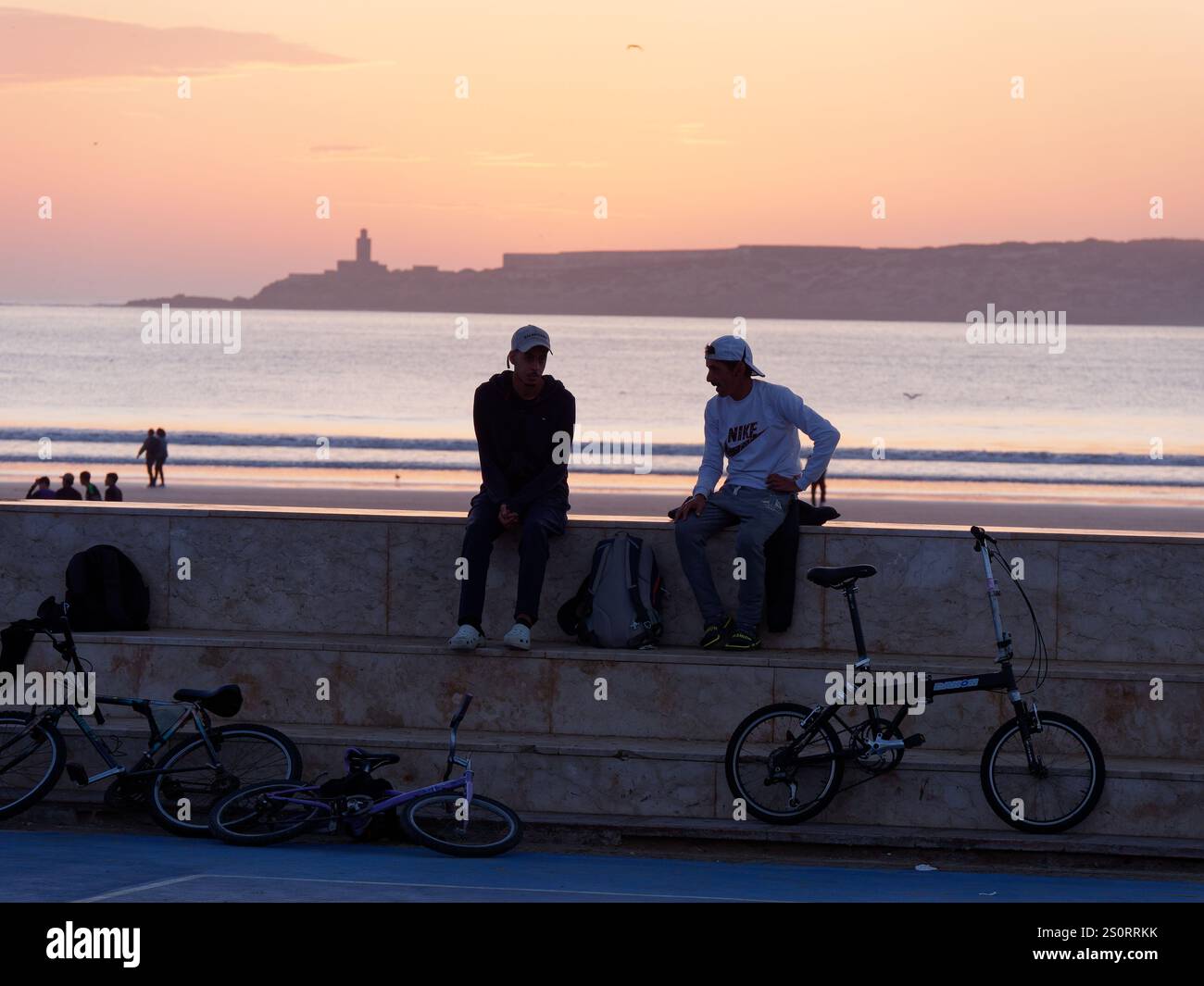 Tramonto sul mare Atlantico e sull'isola all'orizzonte con giovani uomini seduti su una parete a gradini accanto alle biciclette. Essaouira, Marocco. 28 dicembre 2024. Foto Stock
