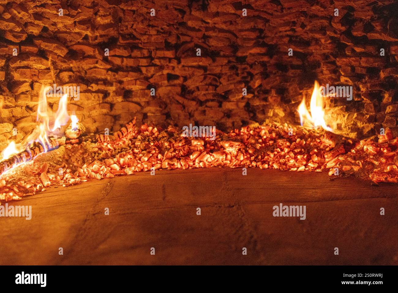 Una vista ravvicinata di carboni luminosi e piccole fiamme all'interno di un tradizionale forno a legna, circondato da un interno in mattoni rustici Foto Stock