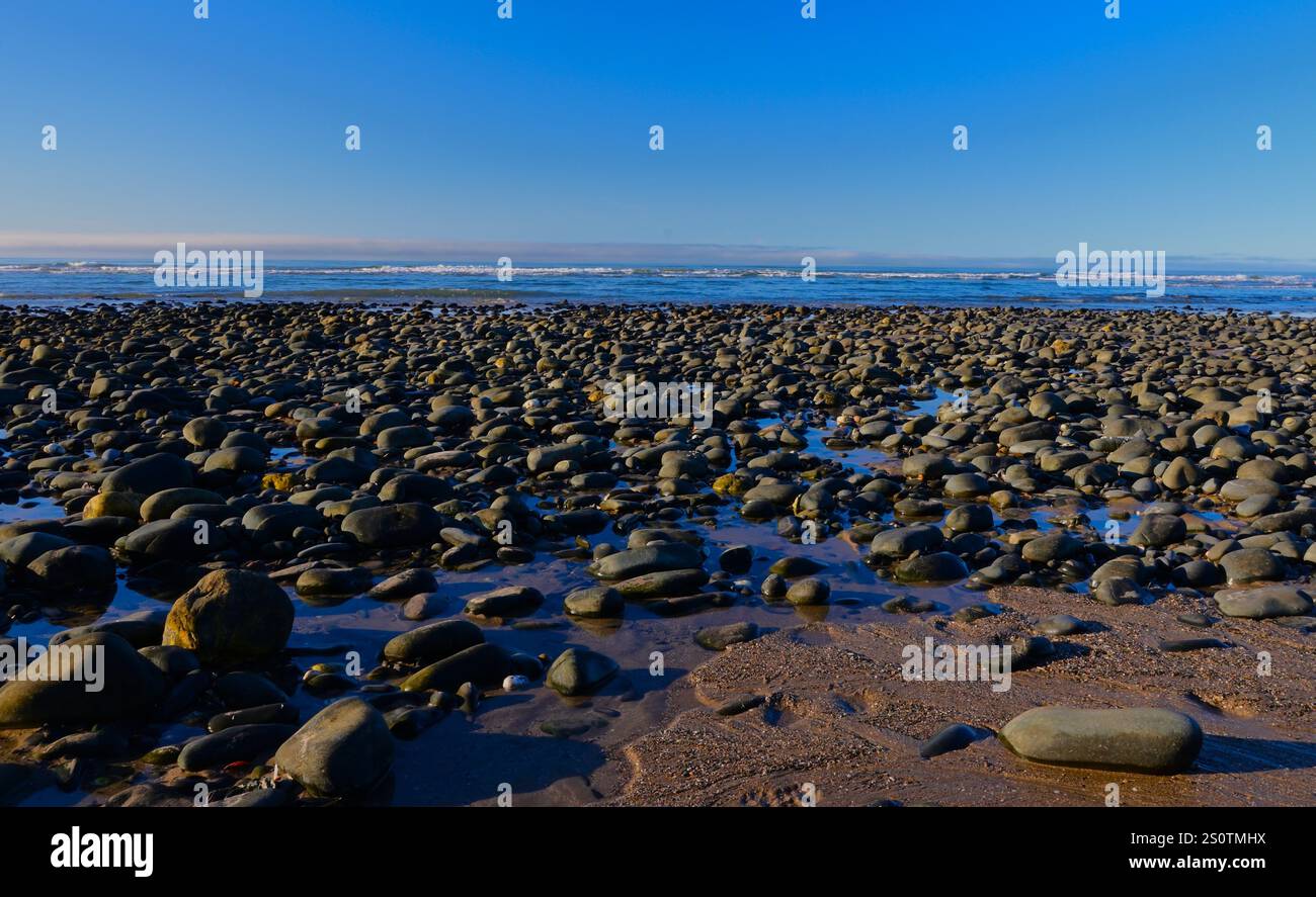 Ciottoli sulla spiaggia, grandi e piccole con piccole onde sullo sfondo sotto un cielo invernale blu. Foto Stock