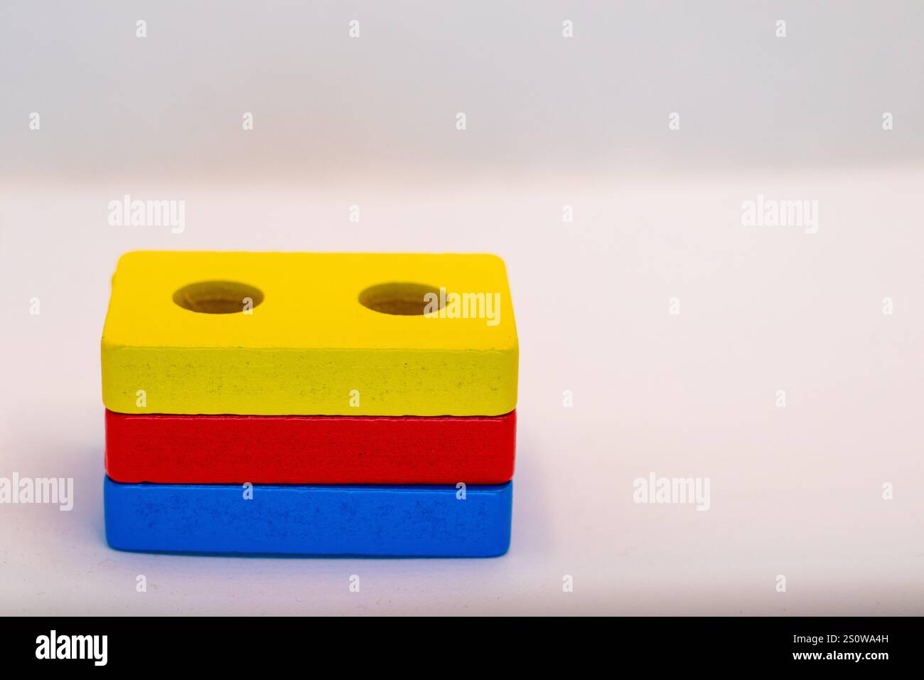 Tre blocchi di legno colorati impilati verticalmente. Il blocco superiore è giallo con due fori, il centro è rosso e il basso è blu. Sono su un whi Foto Stock