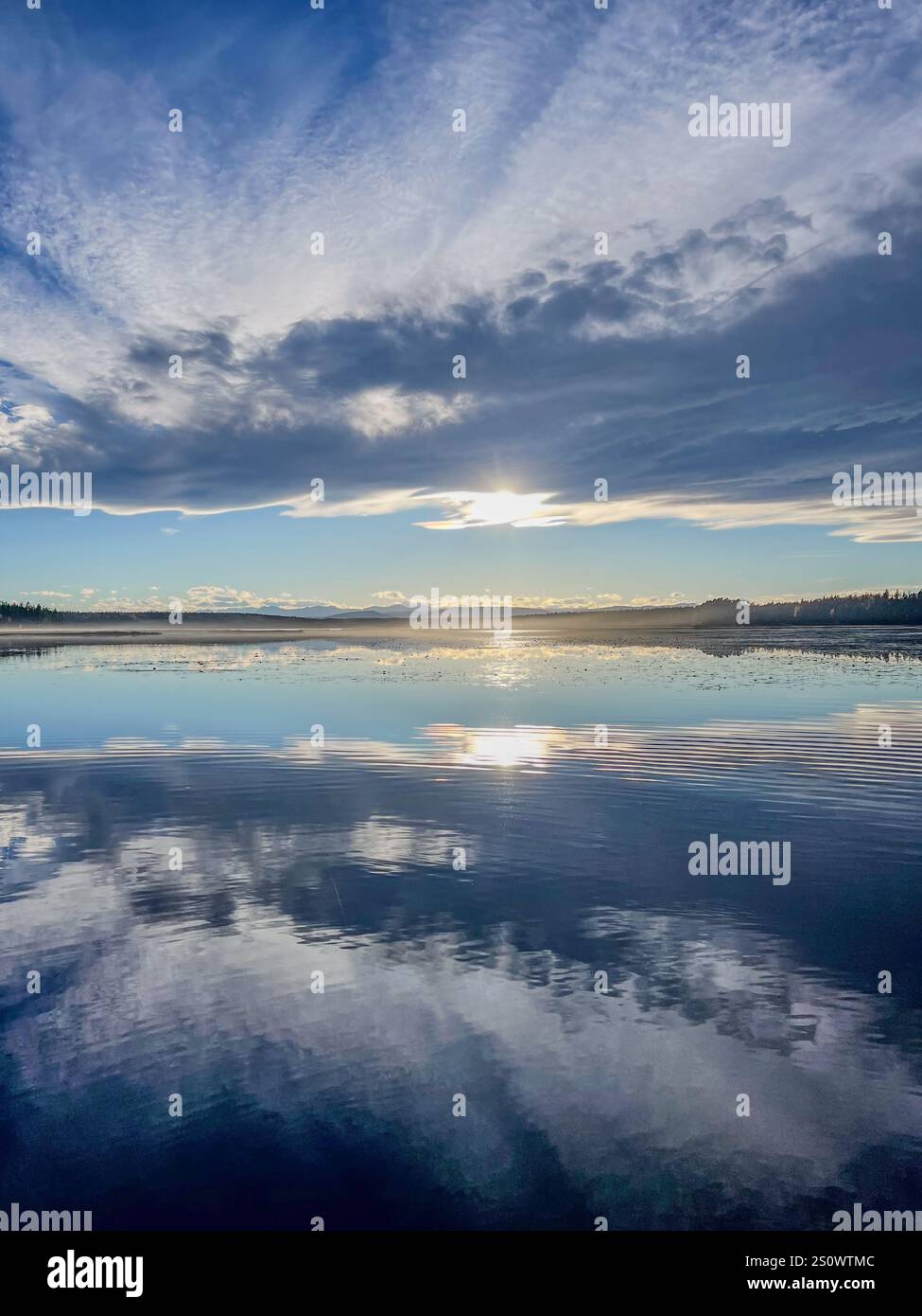 Un lago fermo che riflette un cielo vibrante con la luce del sole che sbircia attraverso le nuvole. Perfetto per paesaggi tranquilli e suggestivi. - Immagine stock catturata con smartphone
