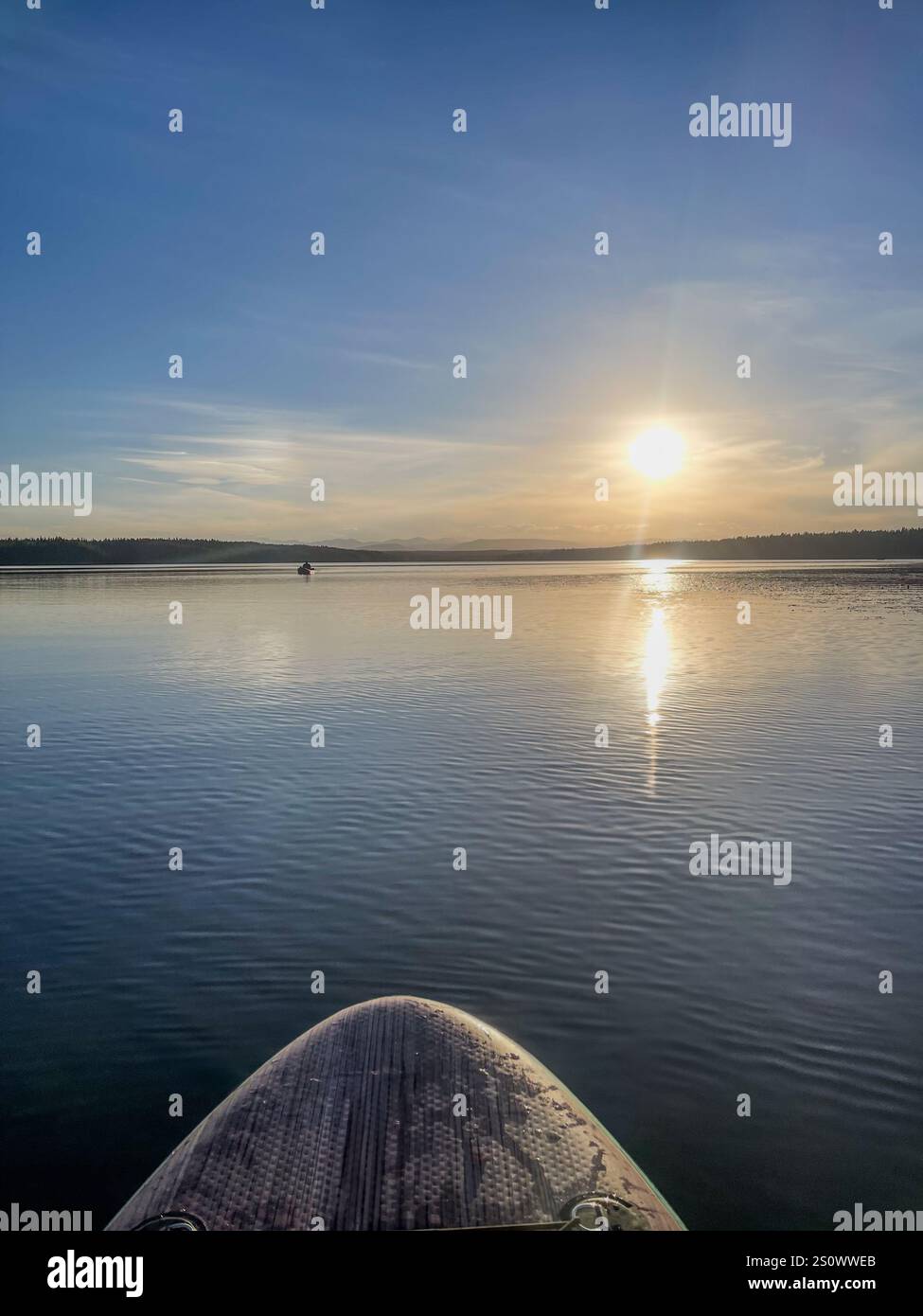 La punta di un paddleboard punta verso il sole che tramonta su un lago tranquillo, con riflessi e montagne in lontananza. Foto Stock