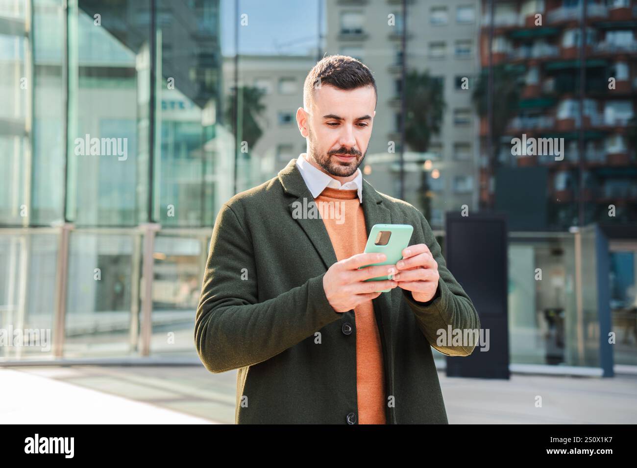 Giovane concentrato che usa un cellulare per inviare messaggi con un'espressione seria mentre si trova all'aperto in centro. Bellissimo uomo che naviga online su Internet con un'applicazione per smartphone. Foto di alta qualità Foto Stock