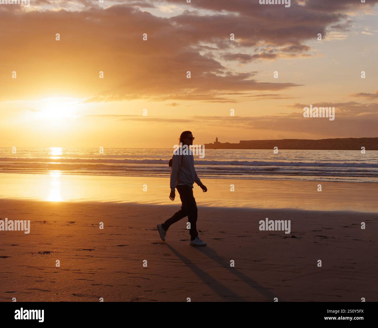 Bel giovane con occhiali da sole e lunghi capelli, passeggiate lungo la spiaggia sabbiosa con il mare e l'isola alle spalle al tramonto a Essaouira, 29 dicembre 2024 Foto Stock