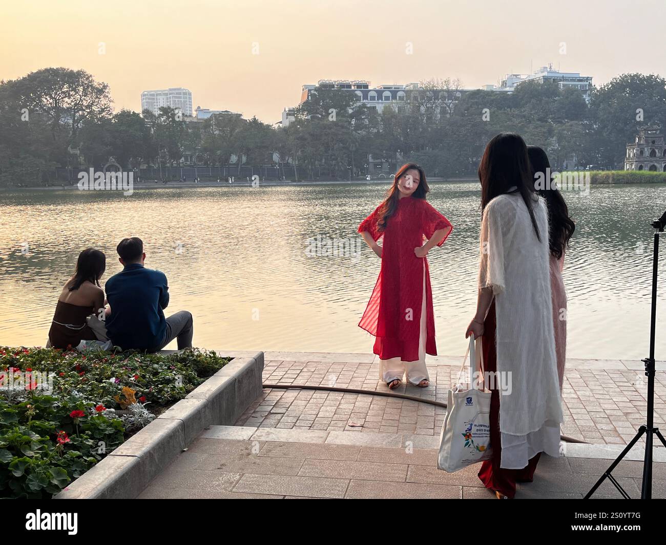 Una donna in abito tradizionale posa per un servizio fotografico al tramonto presso il lago Hoan Kiem, catturando un momento sereno di Hanoi. Foto Stock