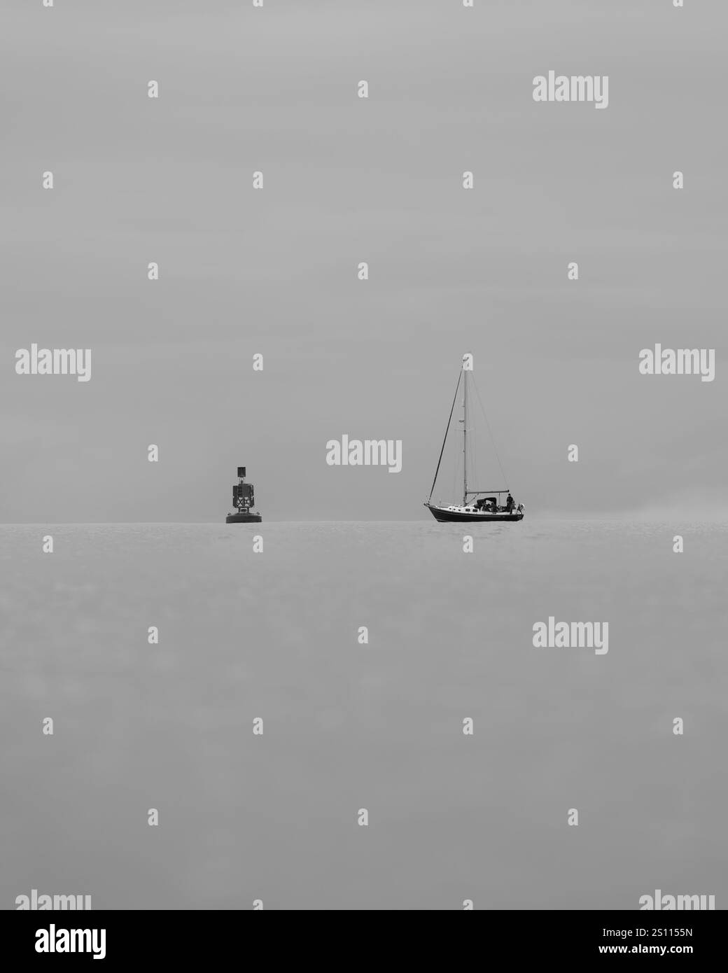 Una barca a vela e una boa riposano su un orizzonte calmo sotto un cielo soffocato e coperto. Il mare è ancora in questa scena minimalista Foto Stock