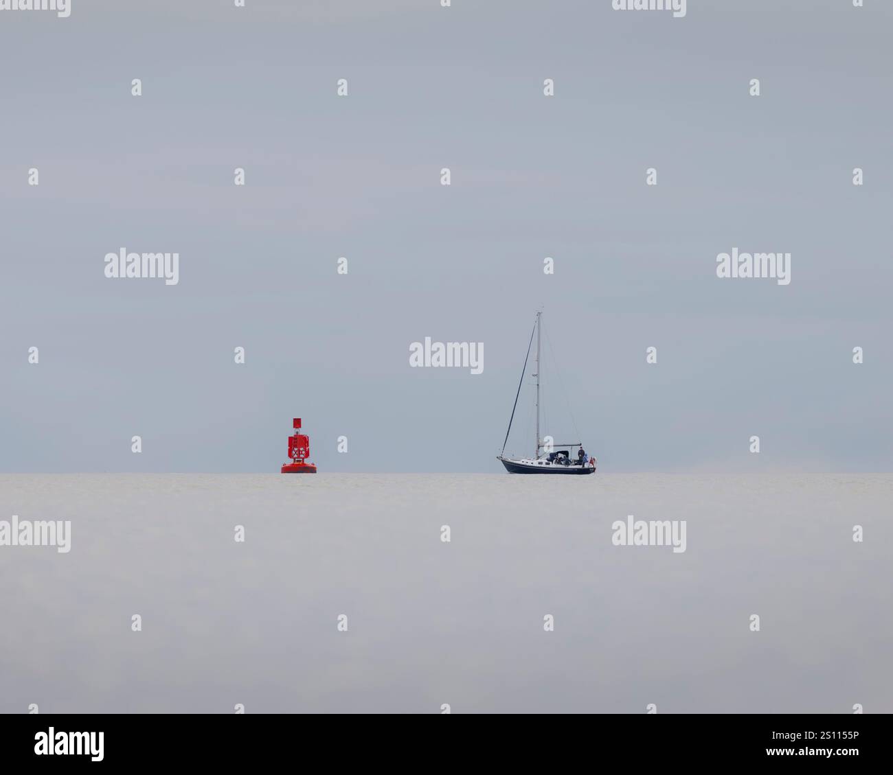 Una barca a vela e una boa riposano su un orizzonte calmo sotto un cielo soffocato e coperto. Il mare fermo e la scena minimalista di Solent, Hampshire, Regno Unito Foto Stock