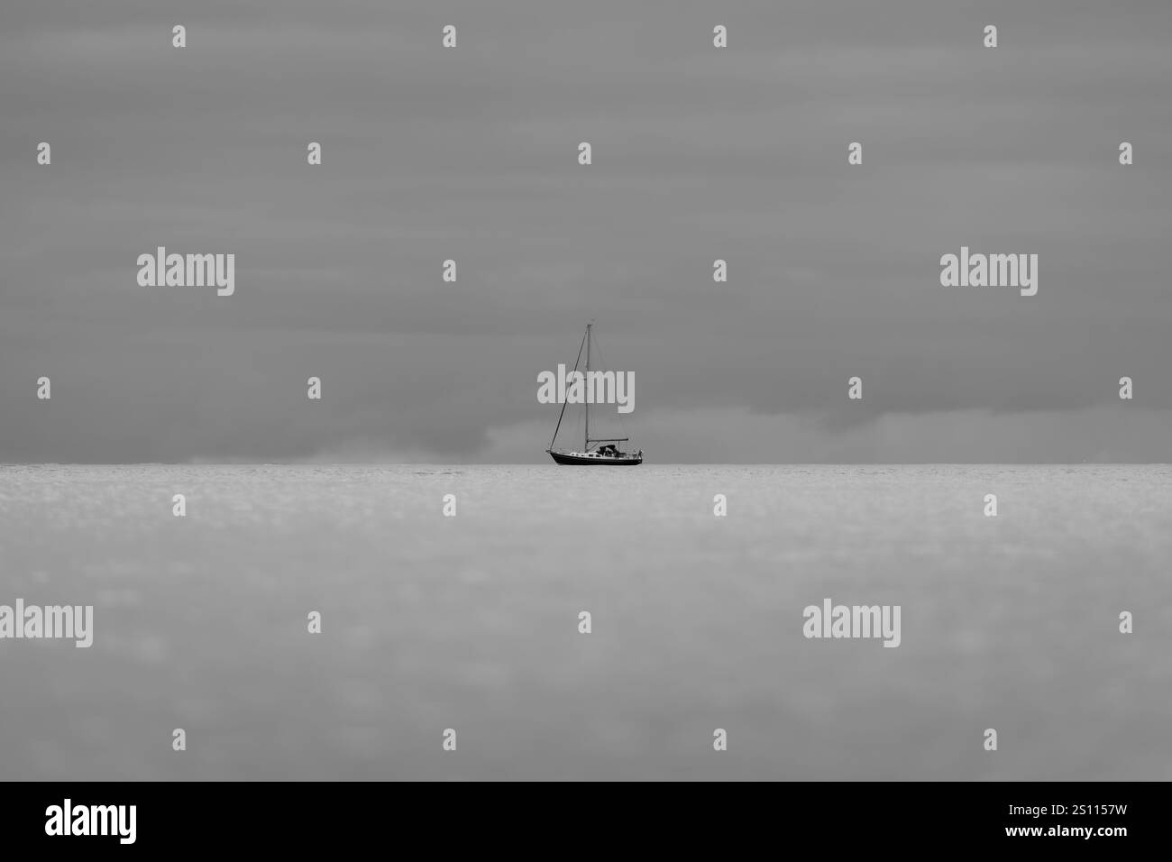 Una barca a vela sembra poggiata su un orizzonte calmo sotto un cielo soffocato e coperto. Il mare è ancora e il cielo scuro in questa scena minimalista. Foto Stock
