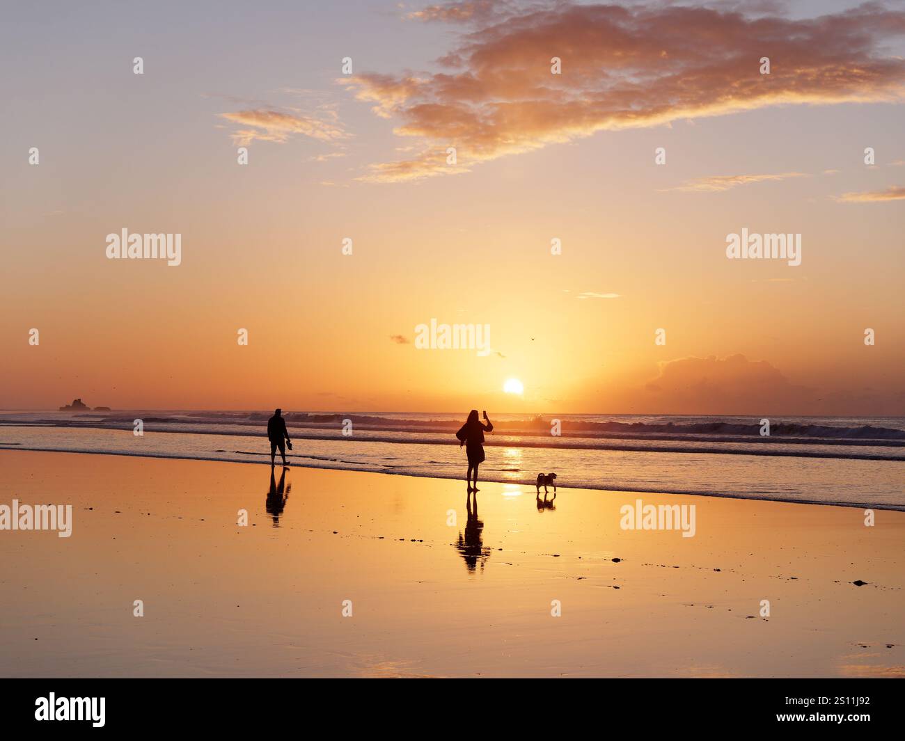 Coppia con un cane che guarda verso il tramonto riflesso nelle acque poco profonde della città di Essaouira, 30 dicembre 2024 Foto Stock