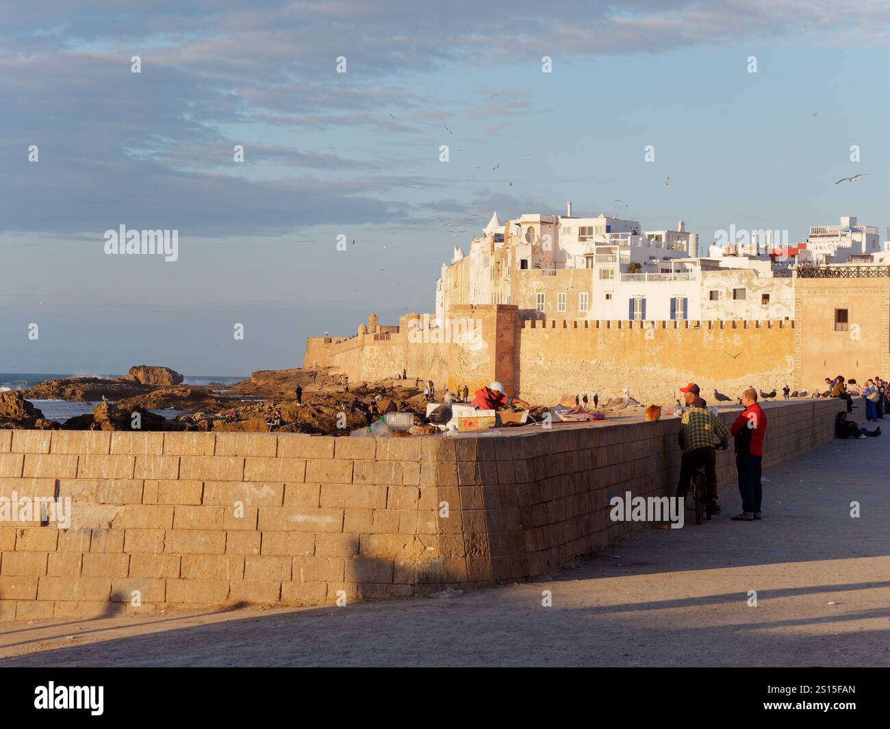 Turisti accanto alle mura della città con la storica Medina alle spalle e la costa rocciosa lasciata nella città di Essaouira, 31 dicembre 2024 Foto Stock