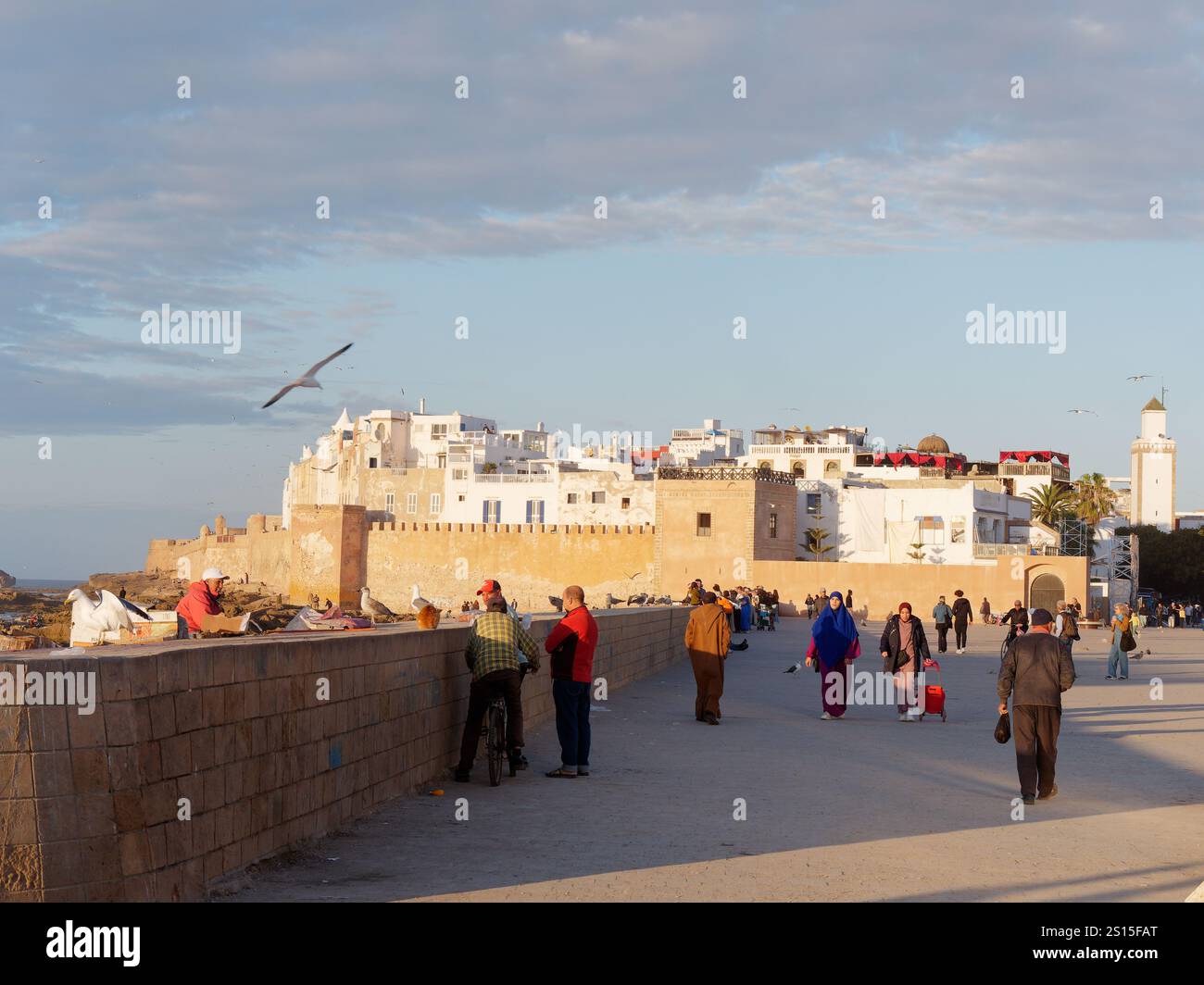 Turisti e gente del posto accanto alle mura della città con la storica Medina alle spalle e la costa rocciosa lasciata nella città di Essaouira, 31 dicembre 2024 Foto Stock