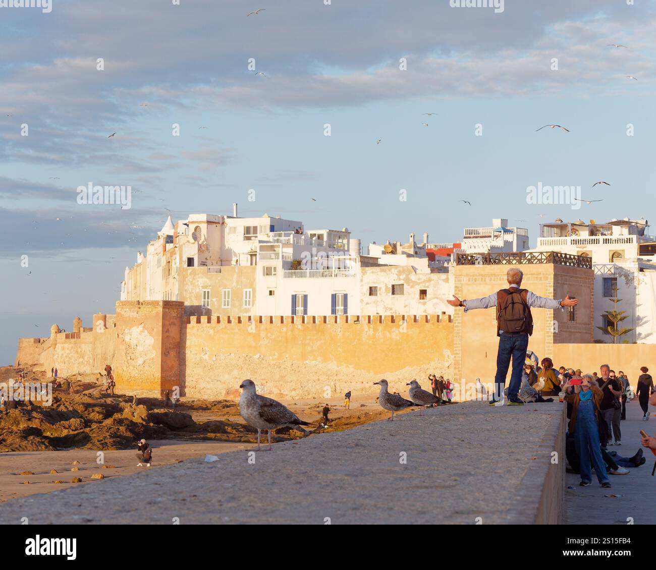 Il turista si distingue sulle mura della città con la storica Medina alle spalle e la costa rocciosa lasciata nella città di Essaouira, il 31 dicembre 2024 Foto Stock