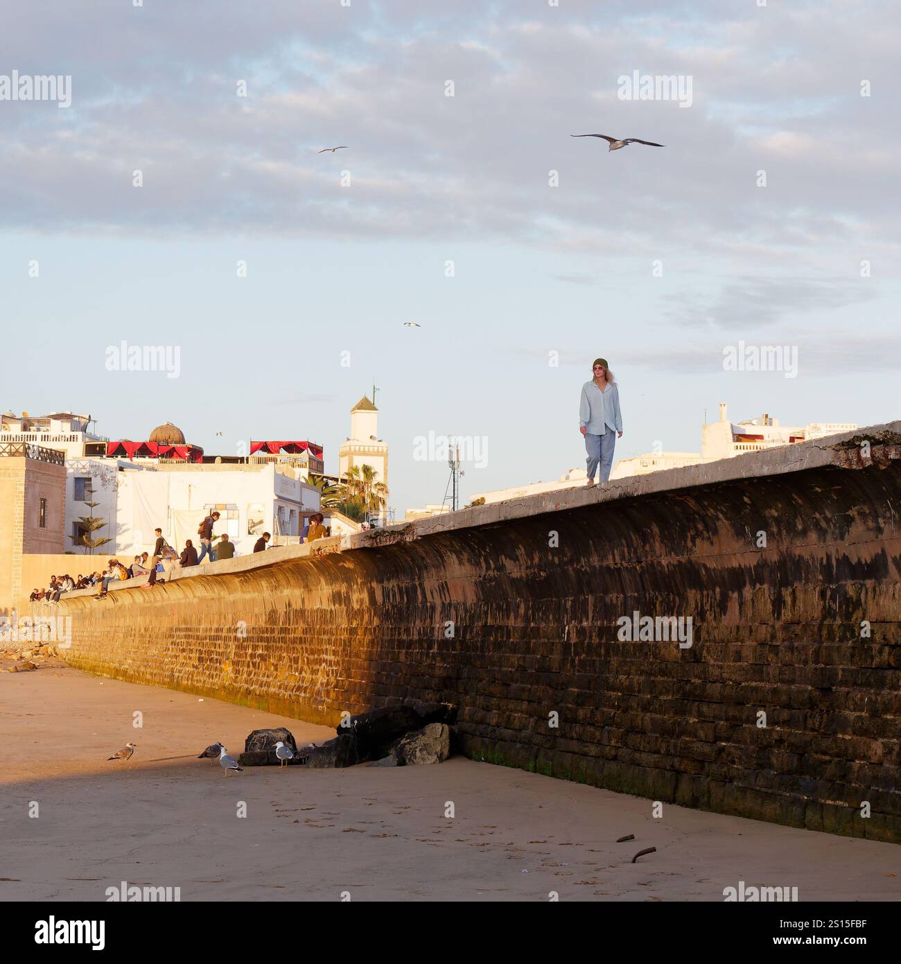 Passeggiate turistiche femminili sulle mura della città con la storica Medina alle spalle e la costa rocciosa lasciata nella città di Essaouira, 31 dicembre 2024 Foto Stock