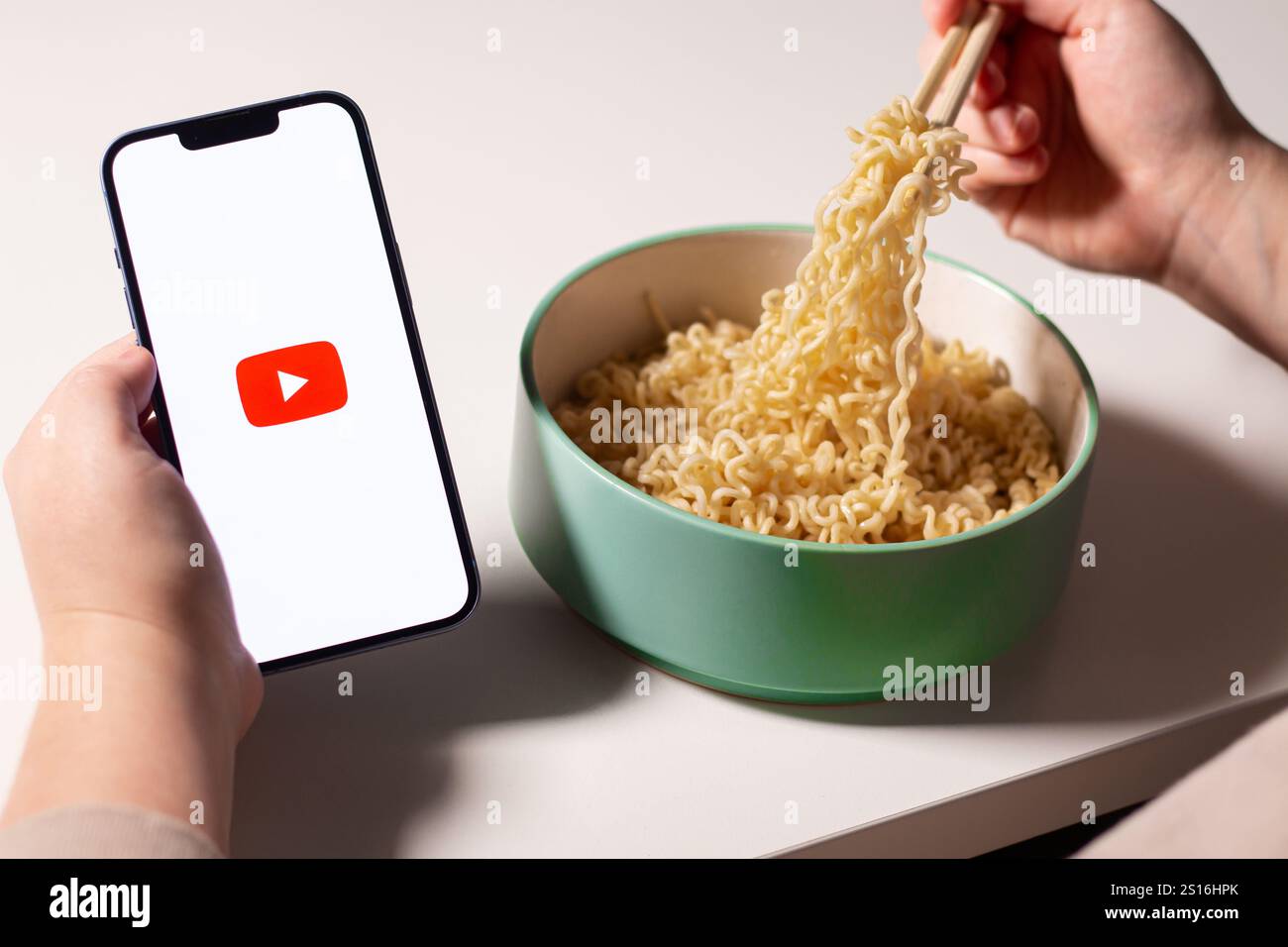 Donna che usa l'app YouTube su smartphone mentre mangia la pasta a casa. Logo YouTube sullo schermo. Mangiare con il concetto di social media Foto Stock