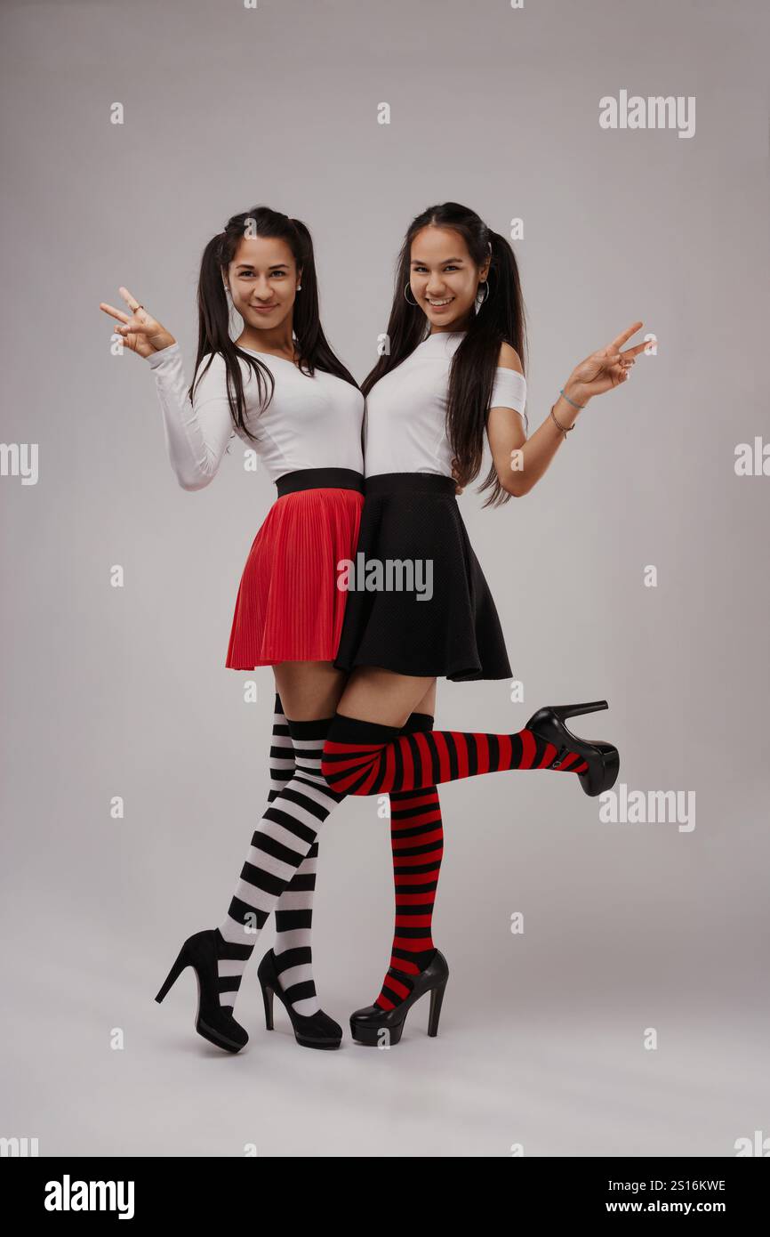 Due giovani donne gioiose che indossano calze al ginocchio vivaci e gonne eleganti si mettono in posa insieme, brillanti segnali di pace e sorrisi irradianti alla telecamera, radia Foto Stock