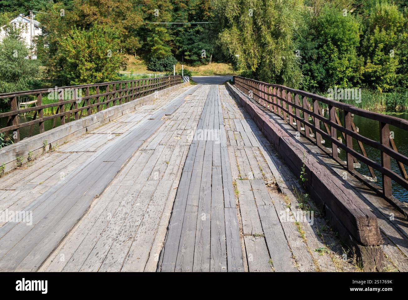 Vecchio ponte di legno con ringhiere su un fiume circondato da lussureggianti alberi verdi. Fotografia prospettica di un paesaggio rurale. Foto Stock