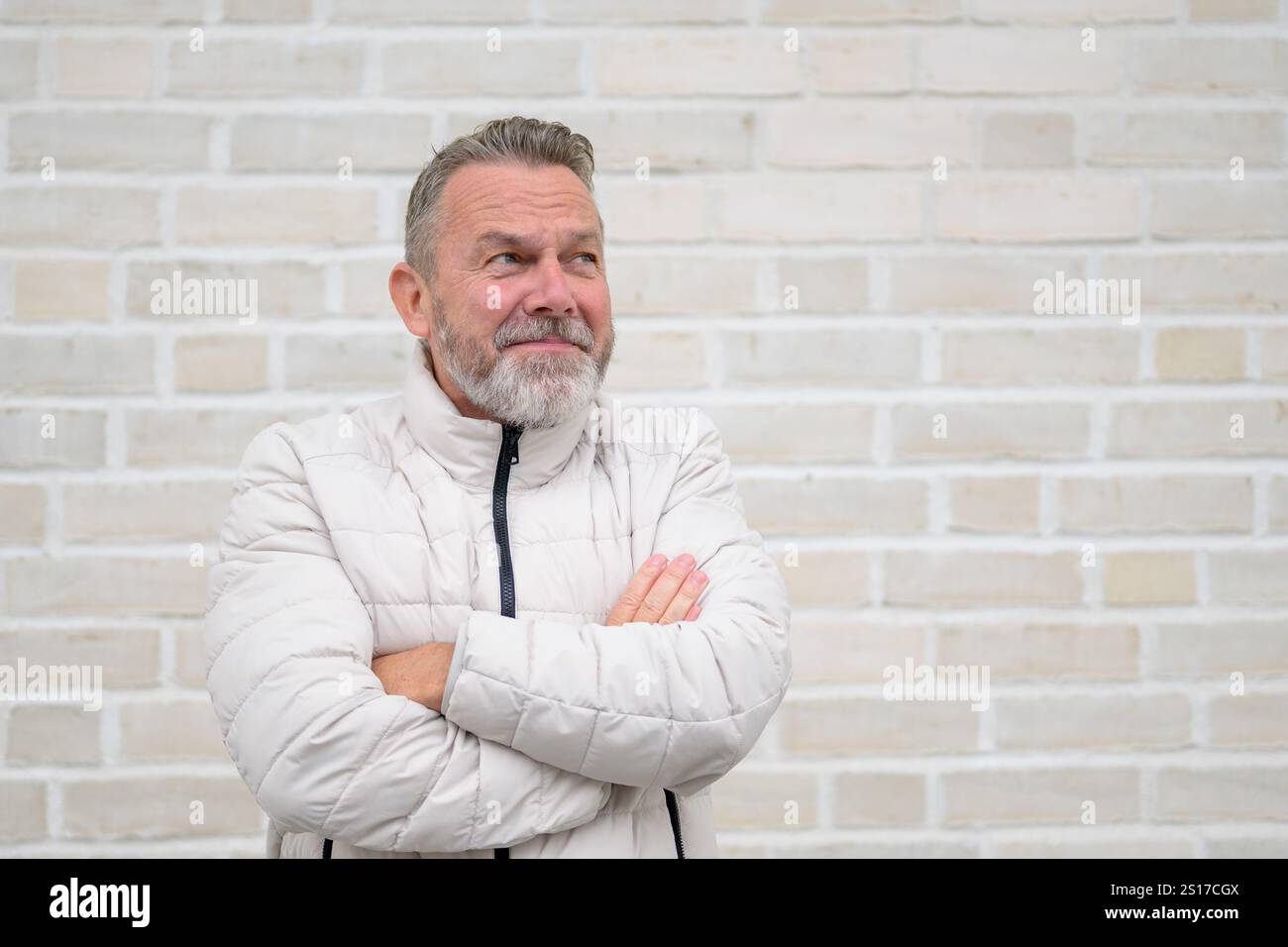 Un uomo maturo con barba grigia e giacca trapuntata di colore chiaro guarda lateralmente con un sorriso malizioso, le braccia incrociate in una postura rilassata. Foto Stock