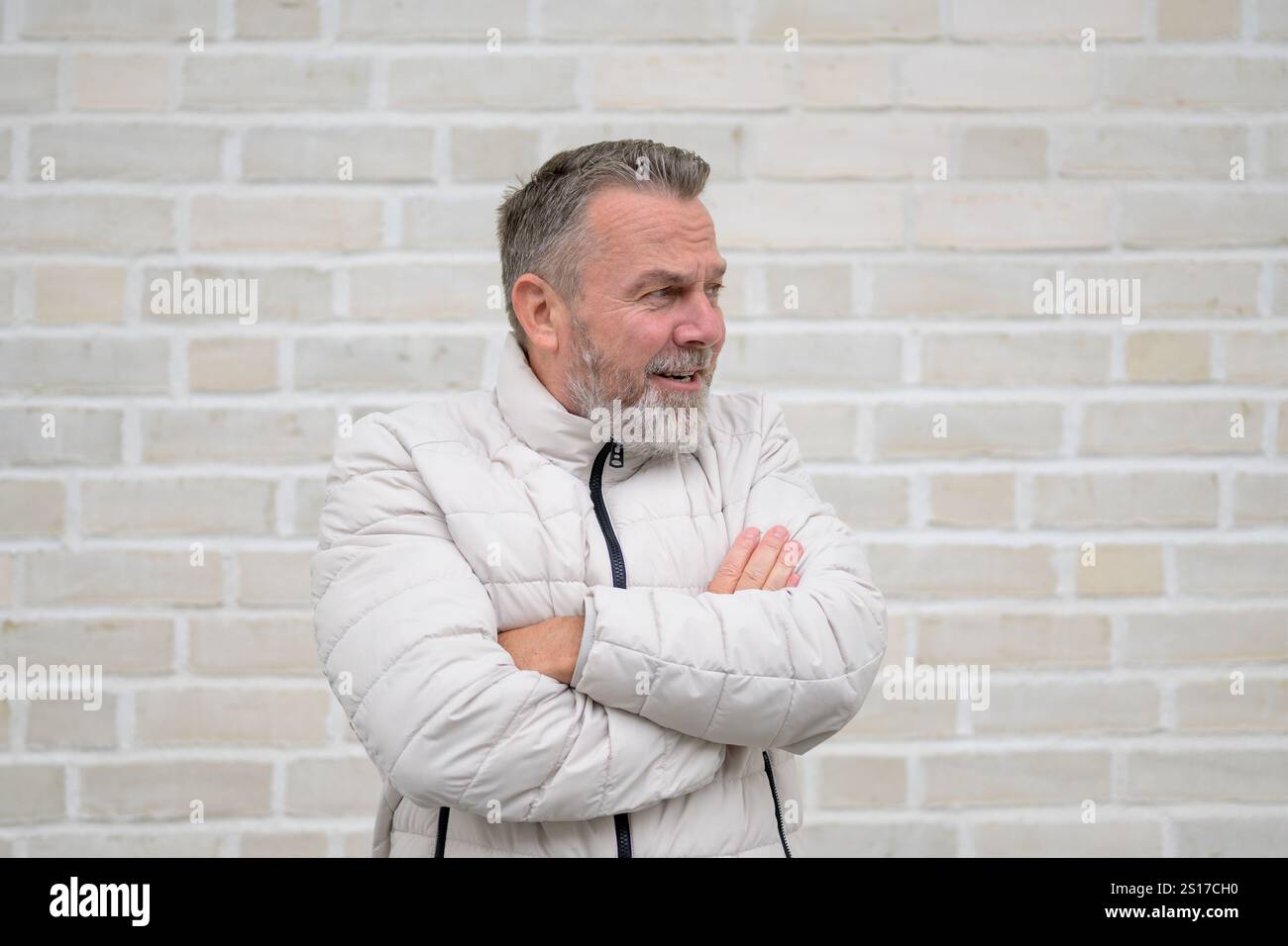 Un uomo maturo con barba grigia e giubbotti trapuntati di colore chiaro con le braccia ripiegate, che guardano attentamente di lato. Foto Stock