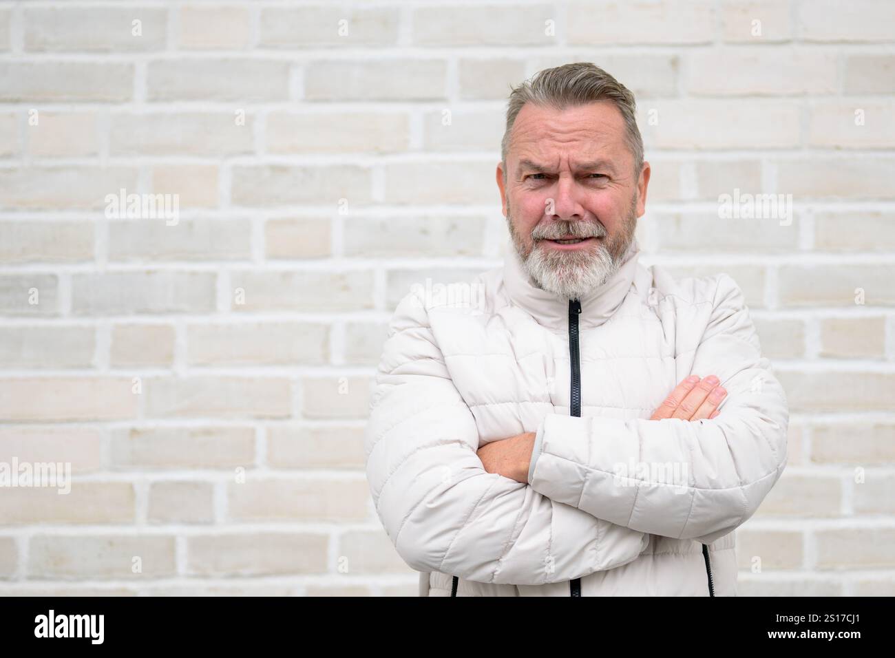 Un uomo maturo con una barba grigia sta in piedi con le braccia piegate, dando un sorriso sottile. Foto Stock