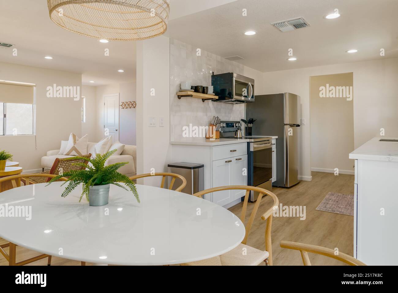 Cucina interna moderna e luminosa con mobili bianchi e pavimento in legno chiaro. Tavolo da pranzo con sedie in legno di vimini. Foto Stock