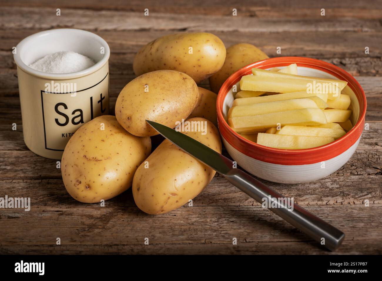 Patate crude accompagnate da una ciotola di patate tagliate alla julienne, un coltello e un barattolo di sale, su un rustico tavolo di legno. Foto Stock
