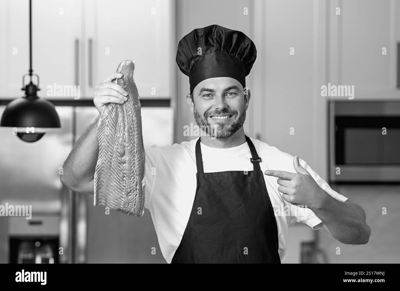 Ritratto dello chef che tiene il salmone di pesce in un cappellino in cucina. Uomo che indossa grembiule e uniforme dello chef e cappello dello chef. Filetto di salmone di pesce crudo per adve Foto Stock