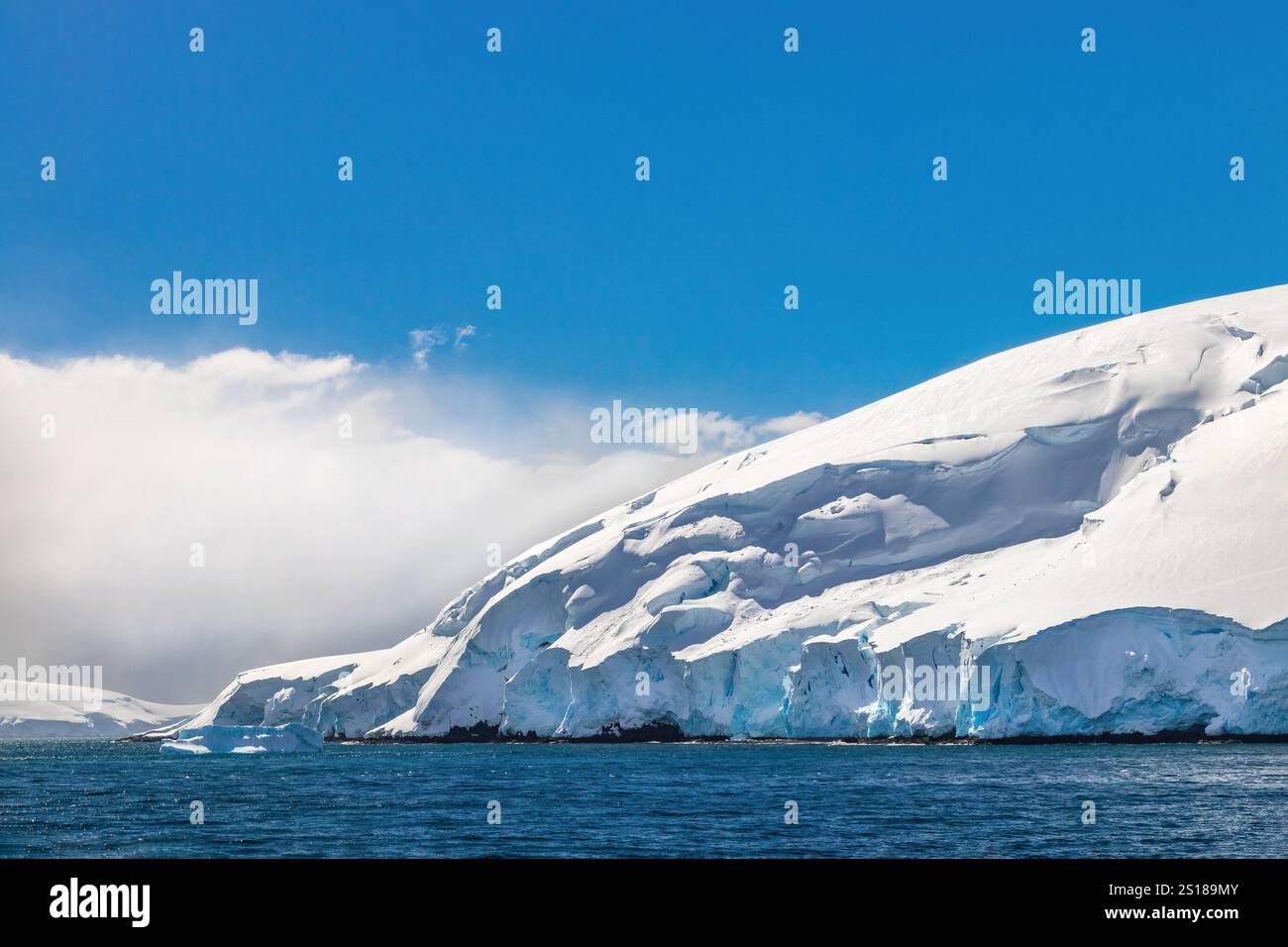 Costa della penisola Antartica coperta di neve profonda. Oceano in primo piano; cielo blu e nuvole dietro. Foto Stock