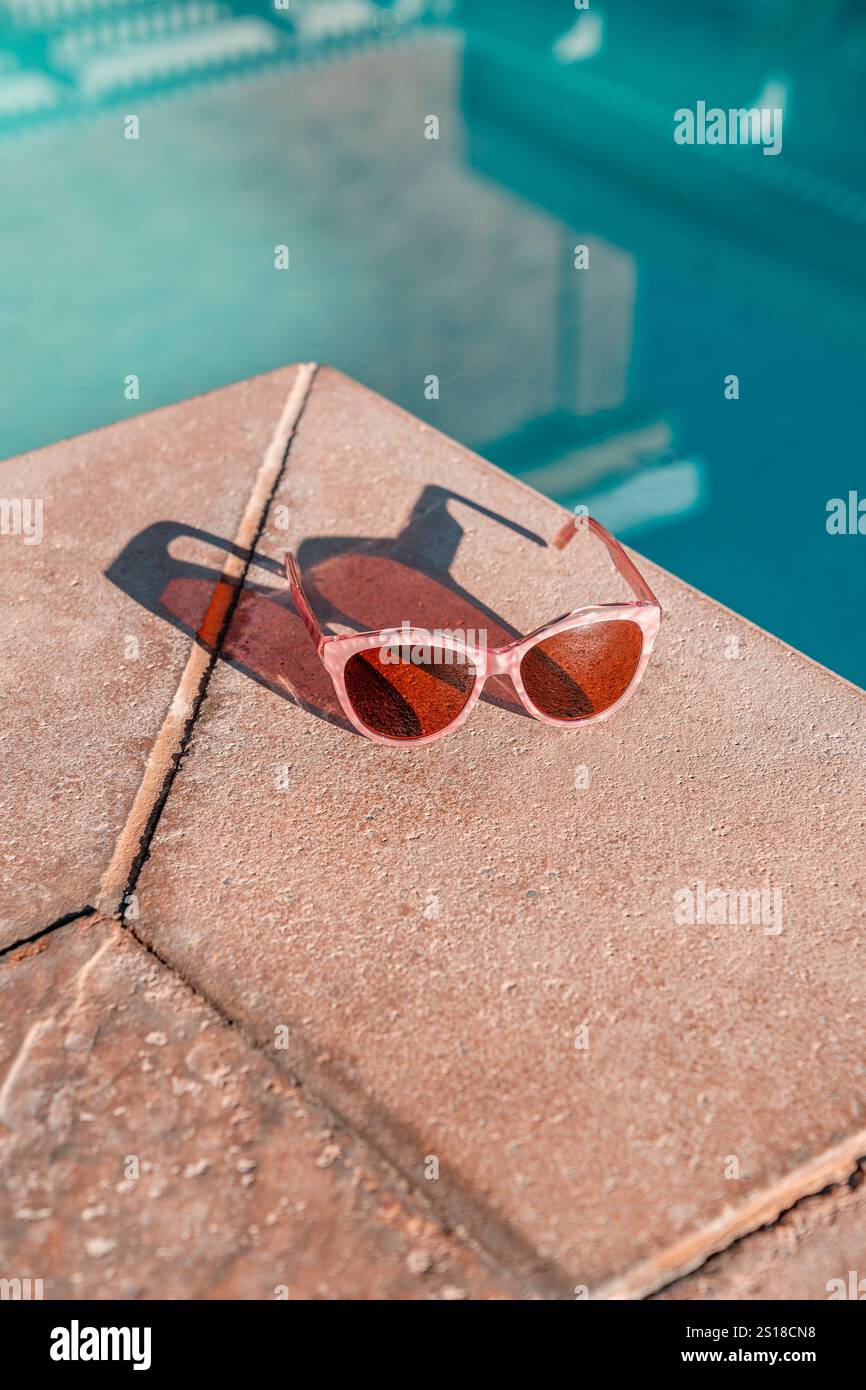 Occhiali da sole da donna con perle rosa sul ponte della piscina a bordo piscina con superficie in pietra ruvida. Piscina blu turchese. Gli occhiali da sole gettano un'ombra. Foto Stock