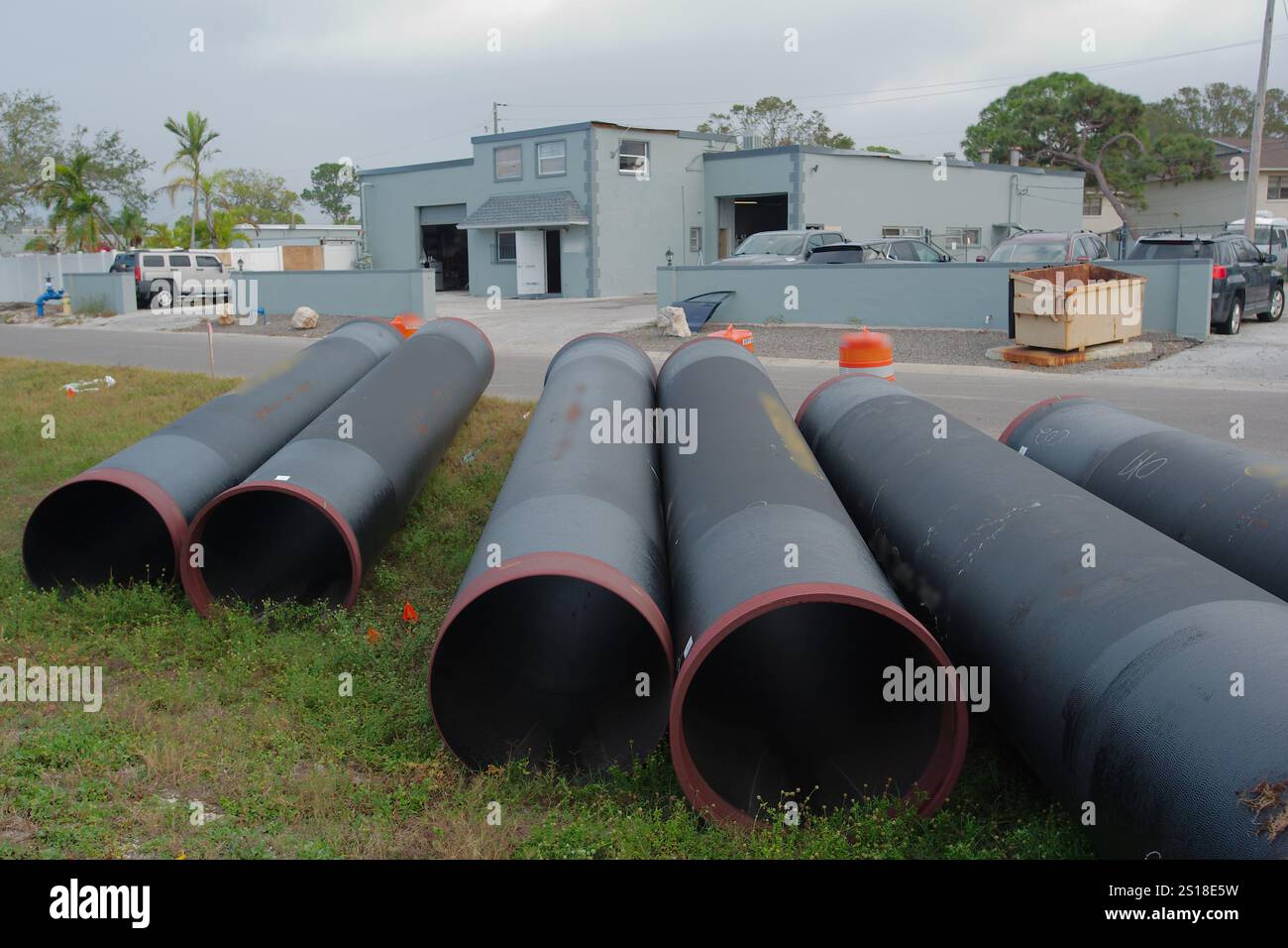 Ampia visuale ribassata su sei tubi principali di trasmissione dell'acqua non trattata neri disposti sul lato della strada e sul bordo dell'erba. Verso un edificio grigio sullo sfondo. Foto Stock