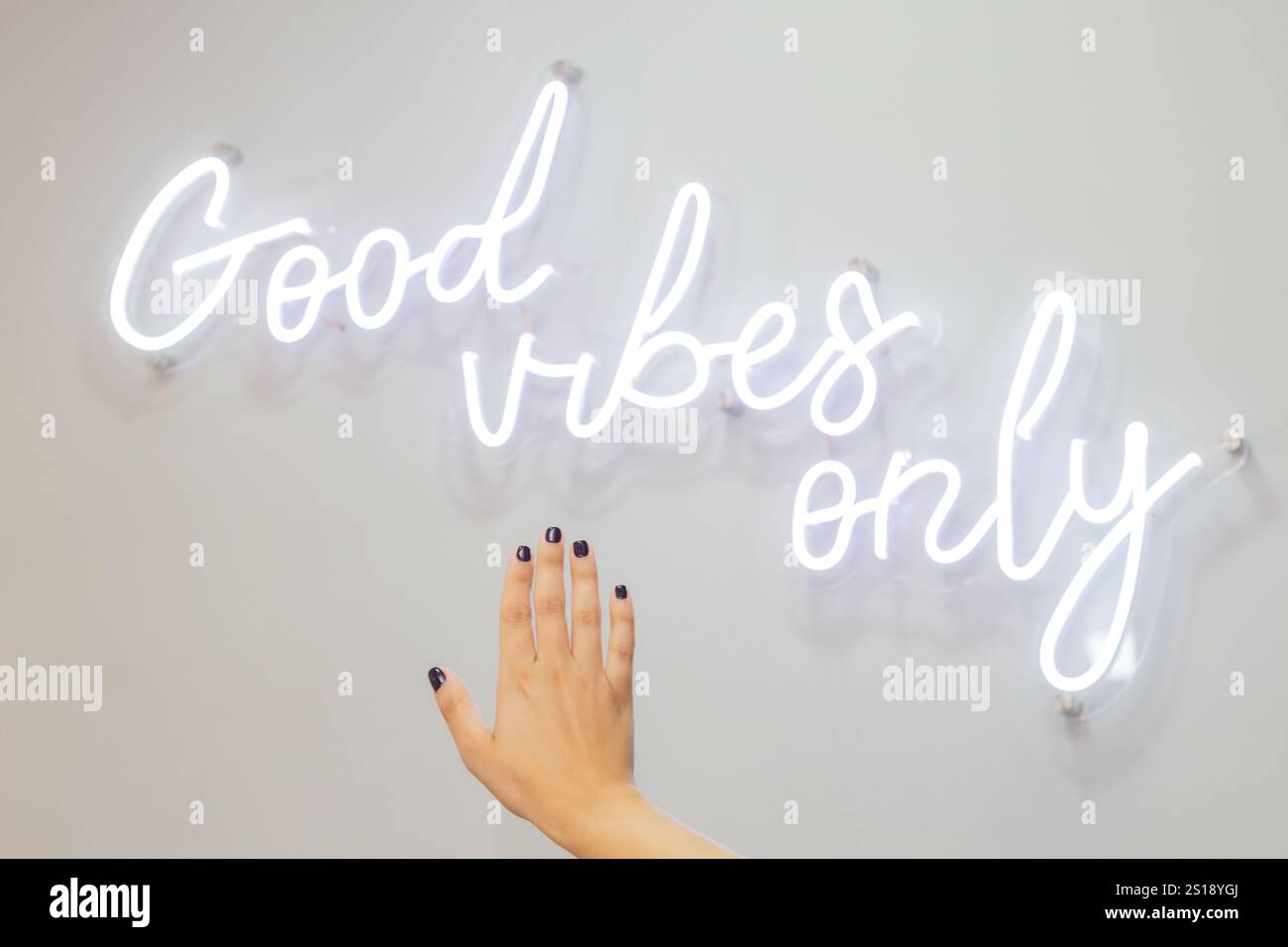 Insegna al neon che recita "solo vibrazioni buone" in bianco brillante, su uno sfondo bianco della parete, una giovane donna mostra le sue mani curate Foto Stock