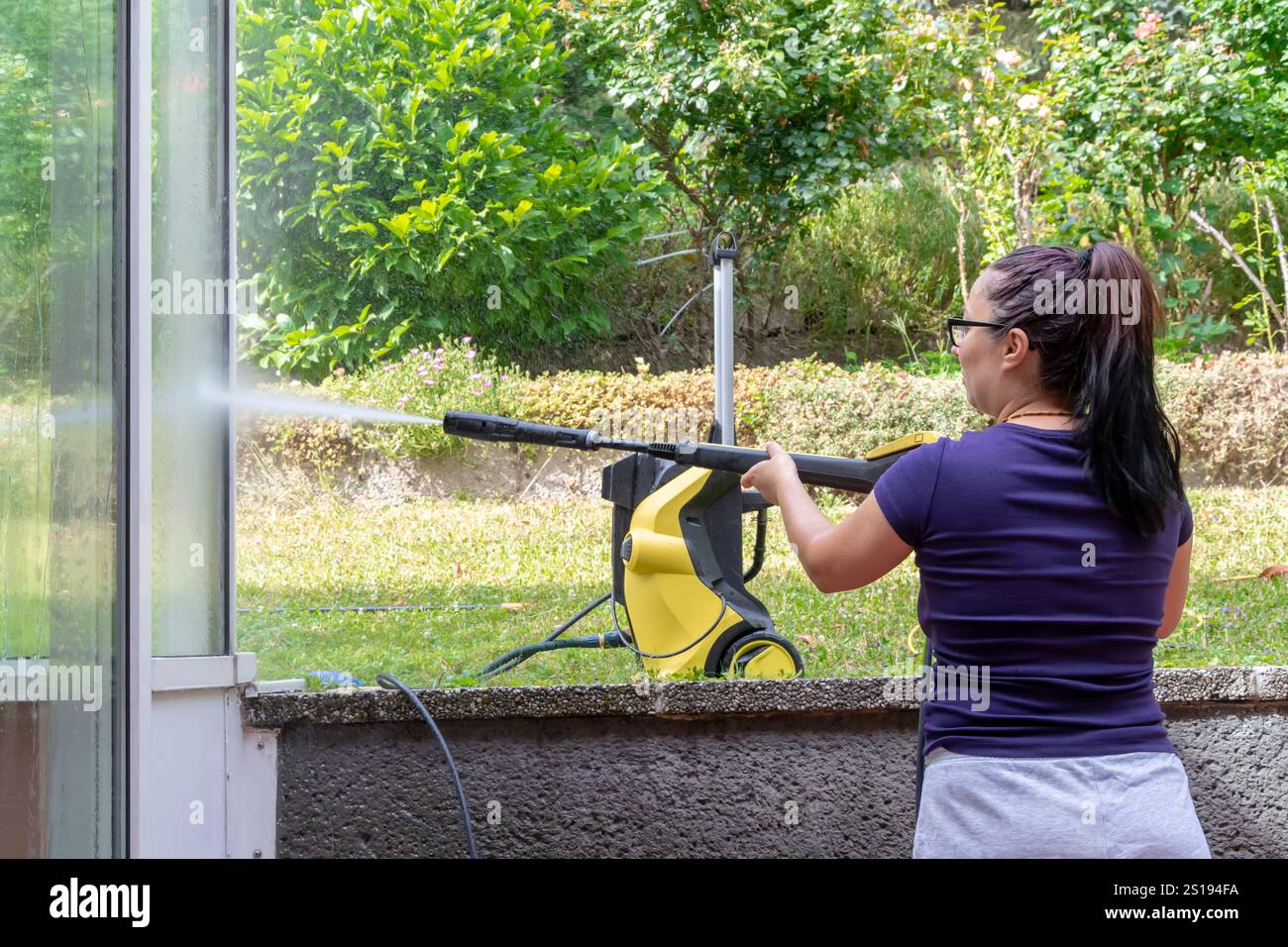Una donna pulisce il vetro a casa con un'idropulitrice. Lavora all'aperto in giardino. Foto Stock