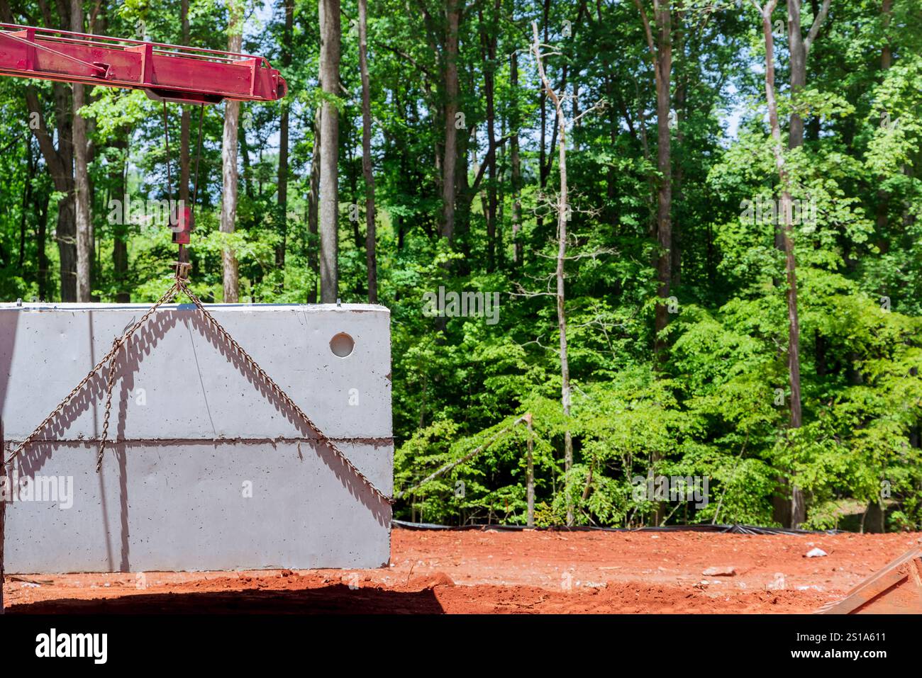 La gru solleva una cisterna settica in cemento sul cantiere circondato da lussureggianti alberi verdi durante la costruzione di una nuova casa Foto Stock