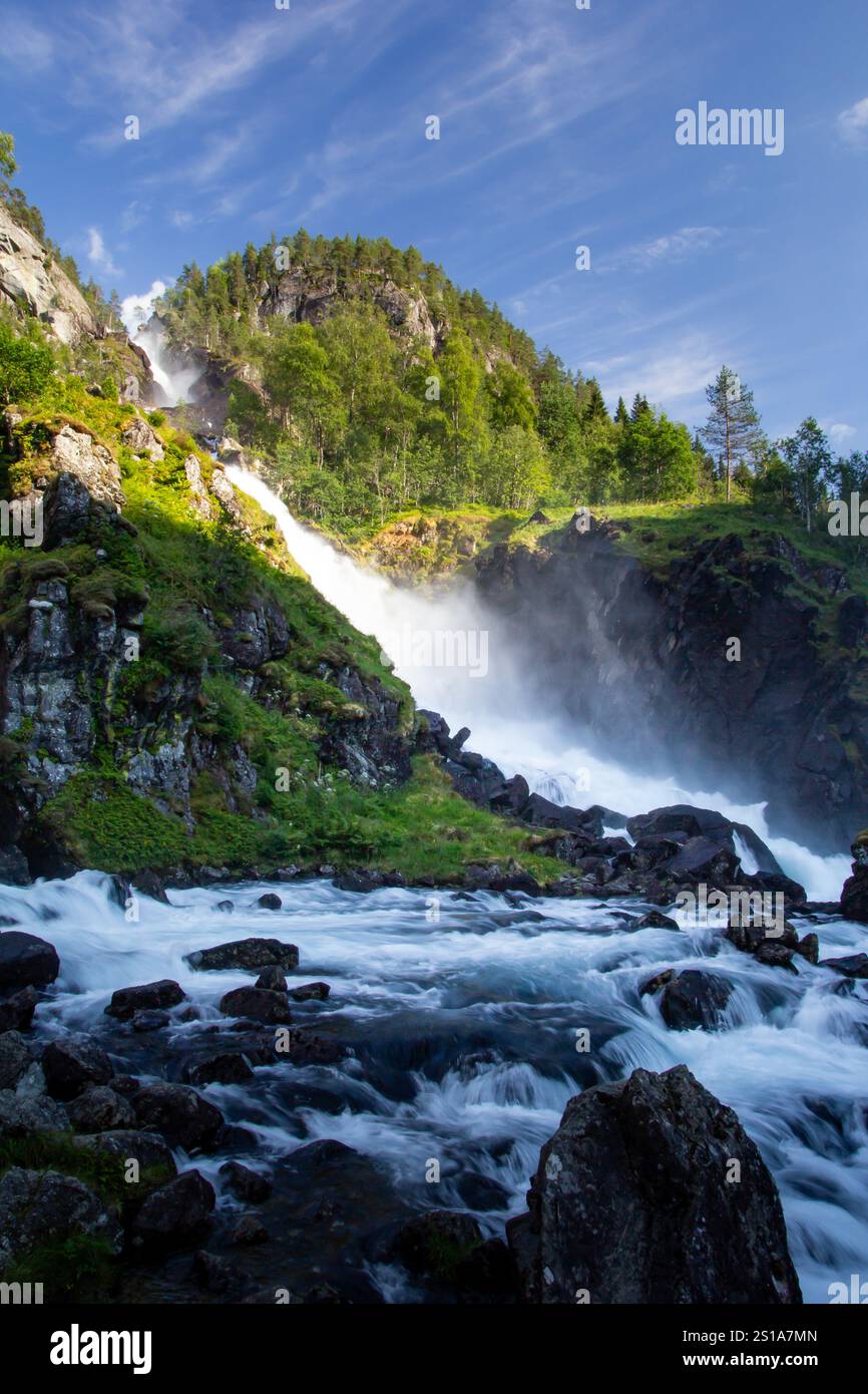 L'acqua scorre sopra una torreggiante cascata, creando nebbia nell'aria, mentre la luce del sole illumina il verde circostante e il terreno roccioso la incornicia. Foto Stock