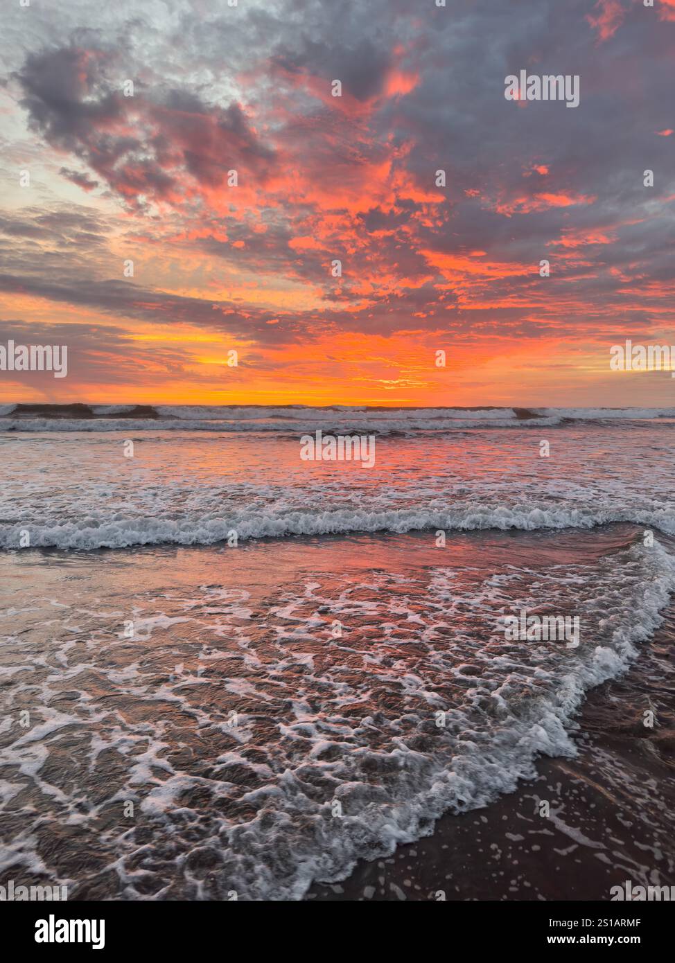 Colori brillanti riempiono il cielo mentre il sole tramonta sull'oceano, creando una vista mozzafiato delle onde che si infrangono a terra. Foto Stock