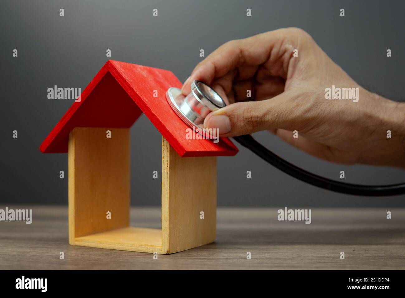 Una mano tiene uno stetoscopio sul tetto di una piccola casa di legno, che rappresenta il concetto di ispezione domestica o di valutazione della proprietà. Foto Stock