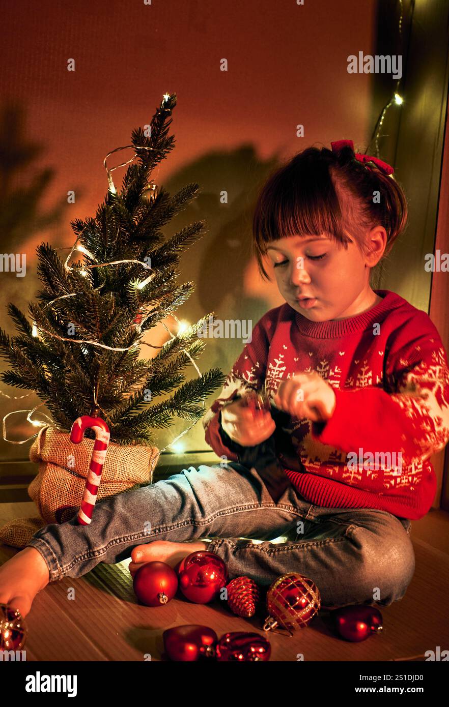 Preparazione per le vacanze e decorazioni natalizie. Una ragazza con un maglione rosso si siede accanto a un piccolo albero di Natale decorato, che regge un ornamento. Bambini in un ambiente accogliente Foto Stock