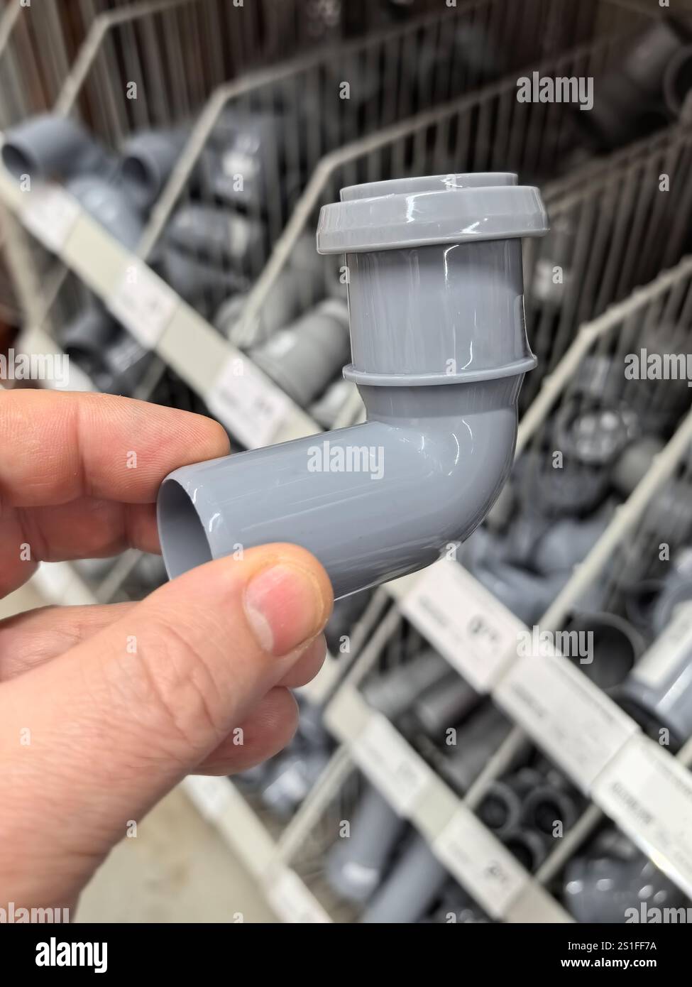 Una mano tiene in mano un gomito grigio per tubi idraulici mentre naviga tra varie forniture idrauliche in un magazzino di ferramenta ben rifornito. Gli scaffali organizzati Foto Stock