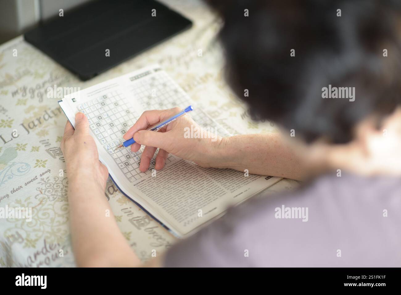 Una donna anziana che risolve un rompicapo con una penna, seduta a un tavolo con una tavoletta sullo sfondo. La scena cattura la messa a fuoco, la concentrazione e. Foto Stock