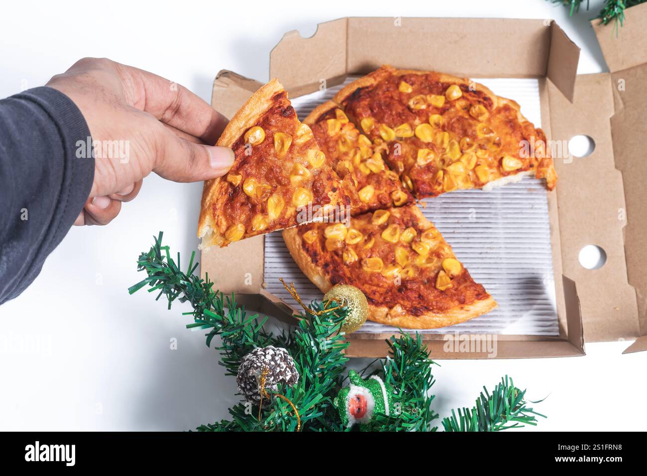Una scena festosa che mostra una mano che prende una fetta di pizza condita con mais da una scatola, circondata da decorazioni natalizie. Perfetto per le vacanze Foto Stock