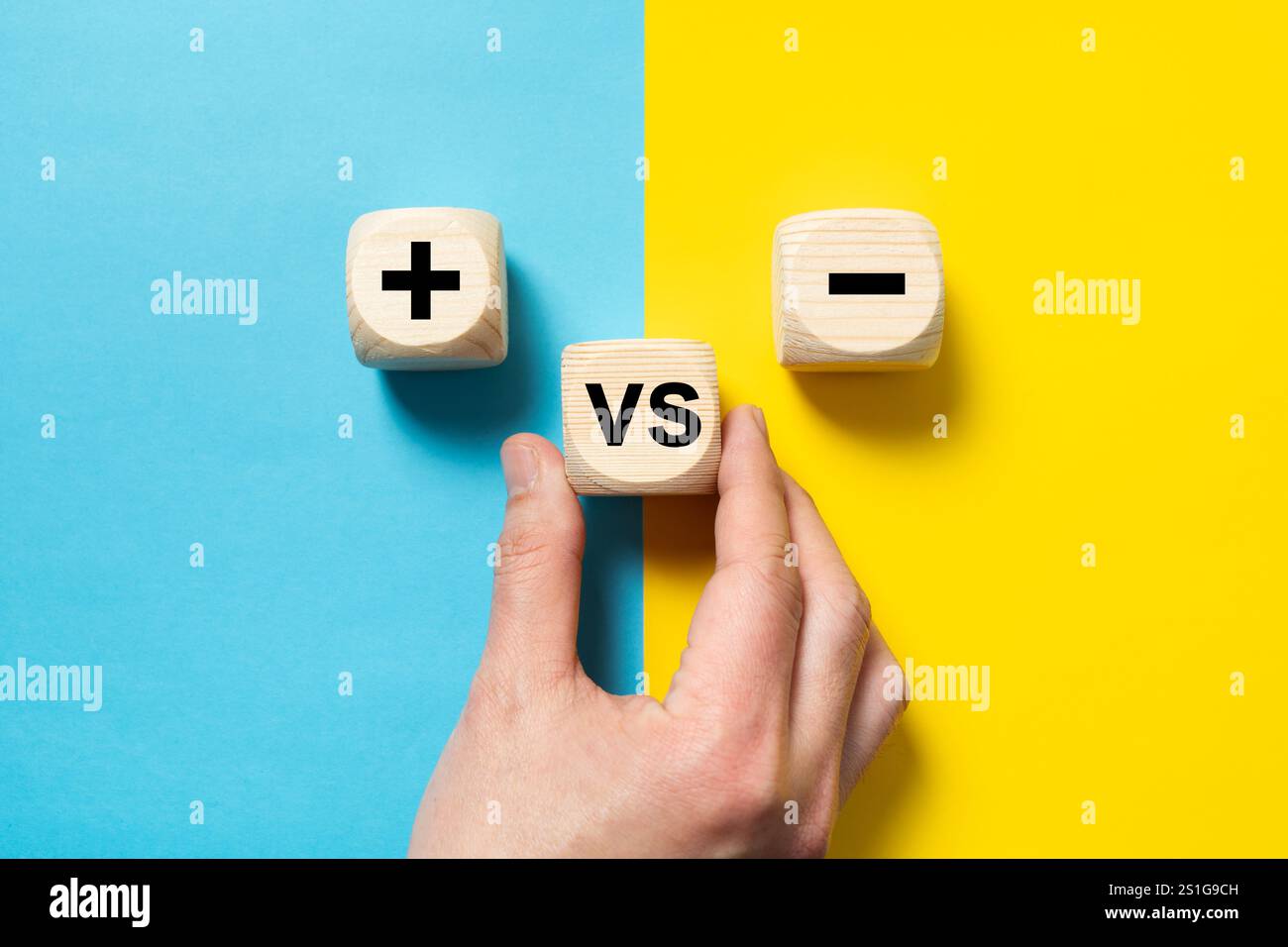 Mano che posiziona un blocco di legno contro il segno più e meno su sfondo blu e giallo, concetto di opposti, pro e contro, posa una composizione piatta Foto Stock