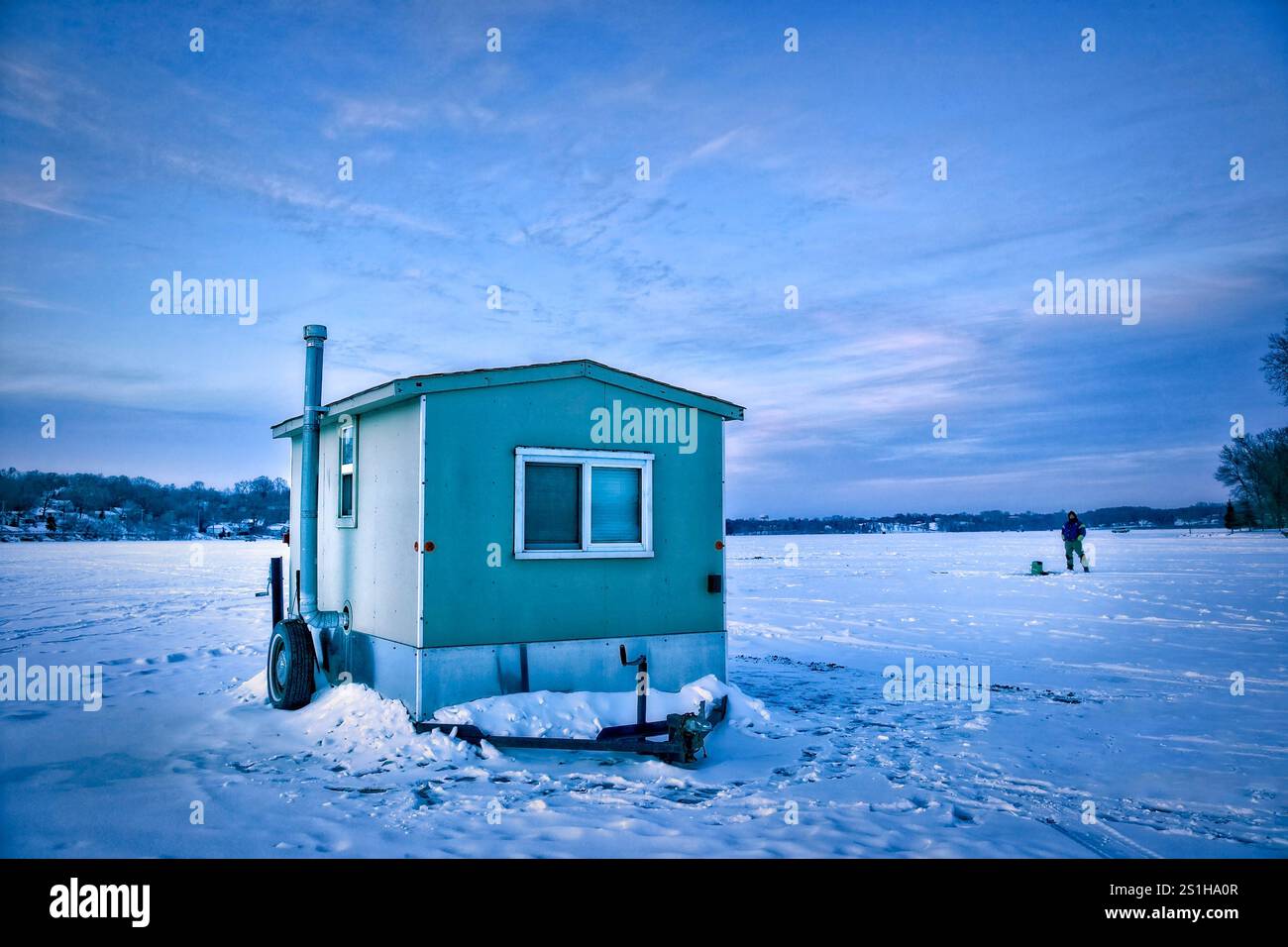 Un pescatore si trova fuori dalla sua rustica baracca per la pesca su ghiaccio su un vasto lago ghiacciato del Minnesota, circondato da un tranquillo paesaggio invernale. L'aria fresca e frizzante Foto Stock