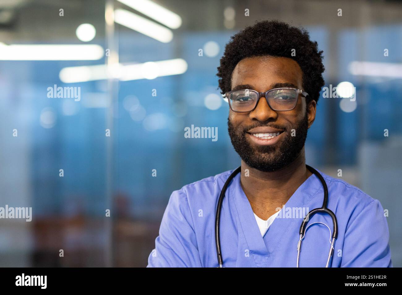 Primo piano ritratto di un giovane medico, l'uomo sorride e guarda la fotocamera. Il medico sta indossando un cappotto medico blu e stetoscopio, lavorando all'interno dell'ufficio della clinica. Foto Stock