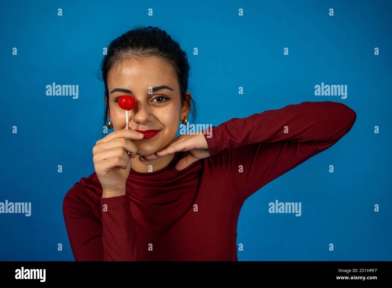Ritratto in studio di una giovane donna allegra che copre un occhio con un lecca lecca, esprimendo gioia e giocosità Foto Stock