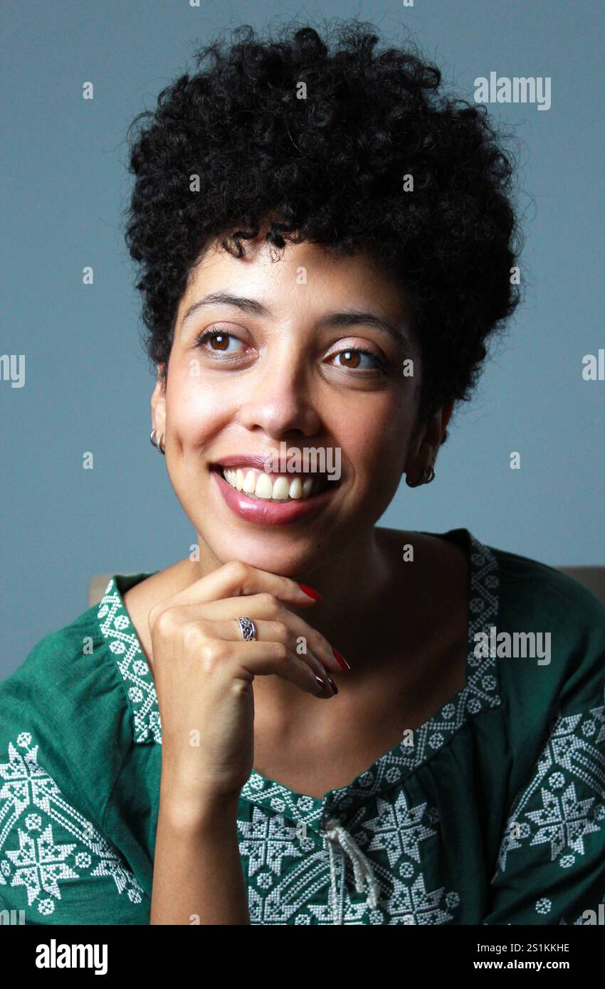 Capelli neri corti che sorridono e sognano ad occhi aperti di fronte a un muro blu. Ritratto verticale della signora afro americana, modello in pelle marrone che indossa una camicetta verde Foto Stock