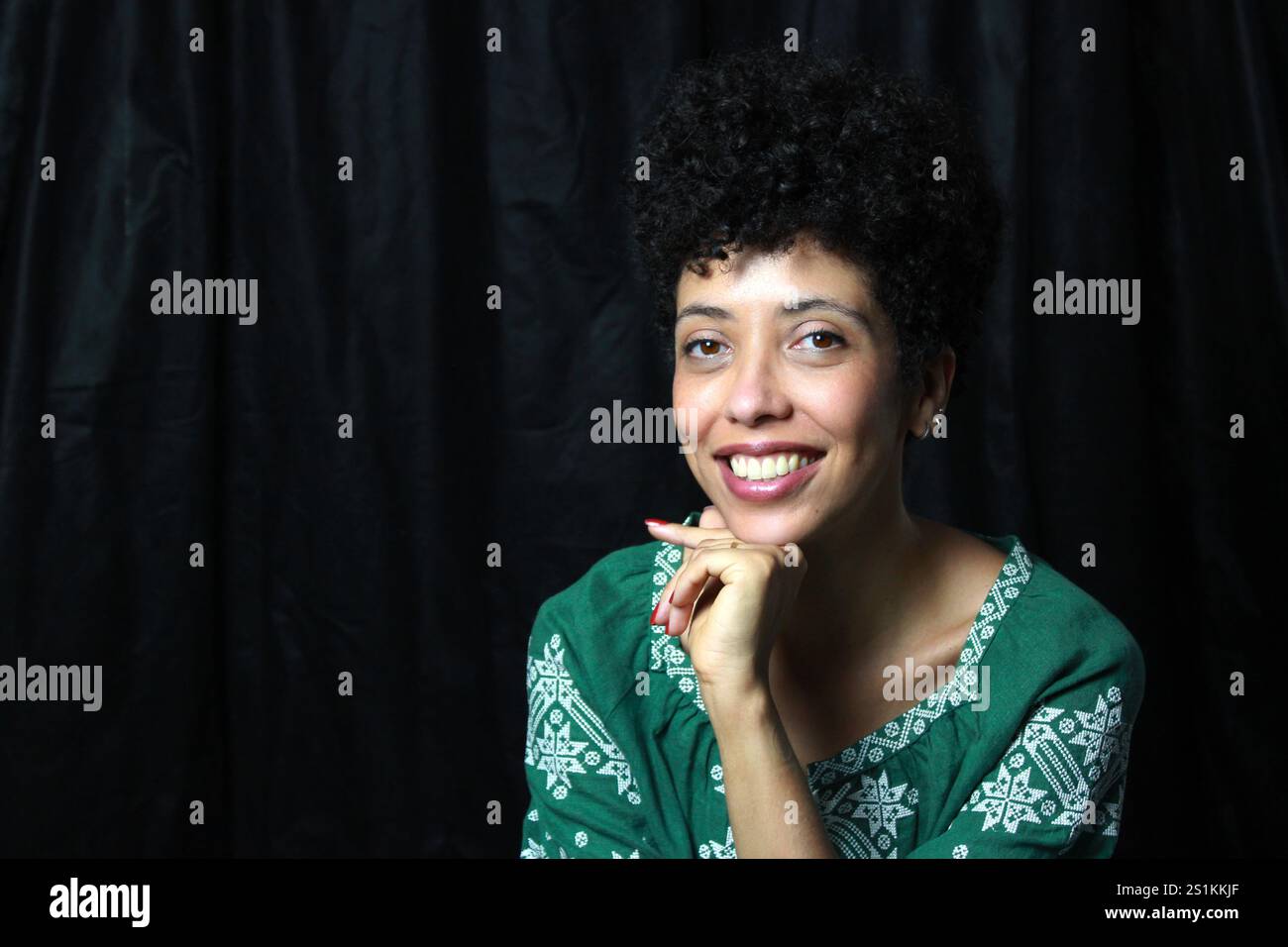 Bella donna dalla pelle marrone che sorride e guarda la macchina fotografica, appoggiando il mento sulla mano. Modello femminile con capelli ricci corti che indossa un abito verde. Foto Stock