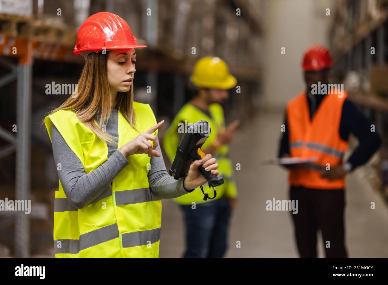 I dipendenti del magazzino che indossano giubbotti di sicurezza e caschi utilizzano la tecnologia per la gestione dell'inventario. Il team collabora in un grande contesto industriale, Foto Stock