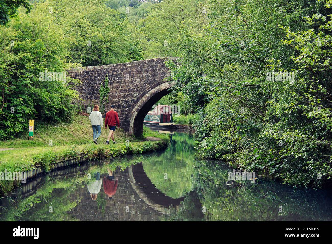 Una coppia che cammina lungo il canale di Caldon vicino al vecchio ponte e si avvicina alla barca stretta con riflessi sull'acqua nelle giornate di sole - vicino a Wetley Rocks, Staffordshire Foto Stock