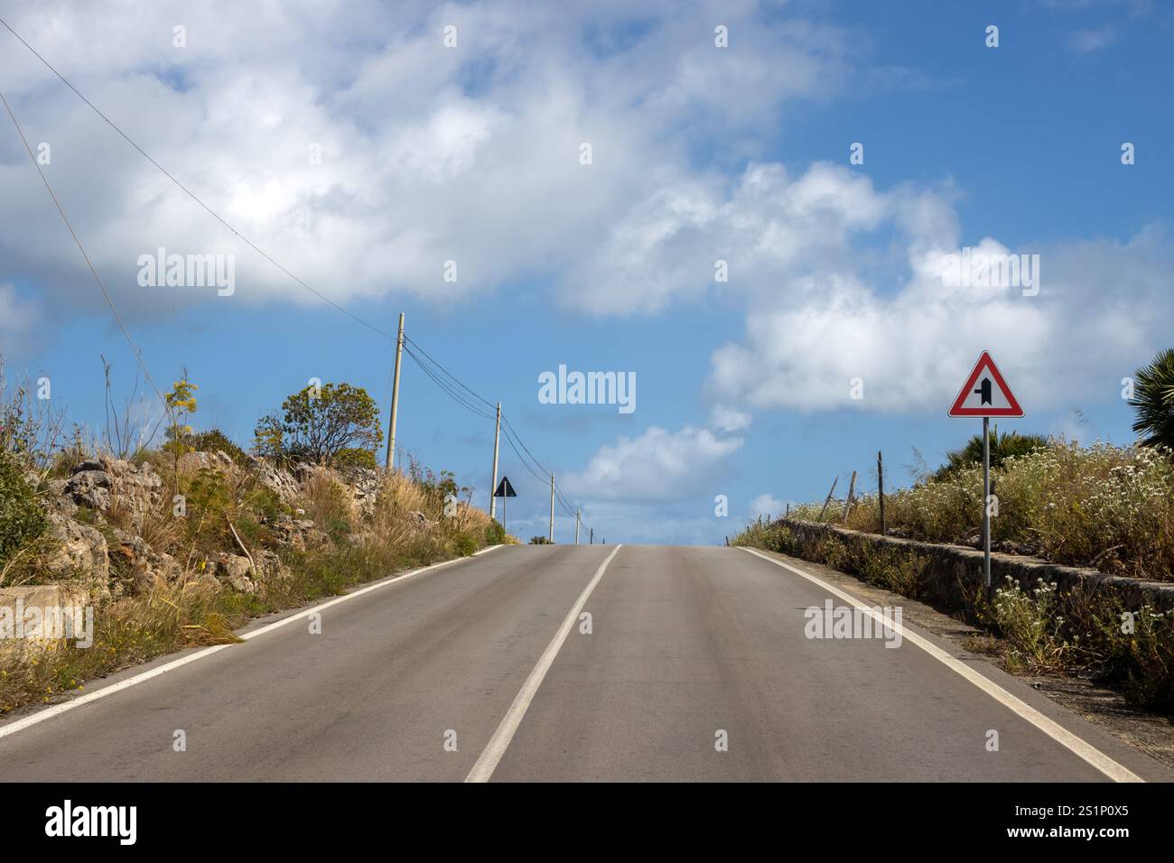 Aspalt Road su una collina, fiancheggiata da cespugli e alberi. Cartello stradale: Incrocio con una strada a sinistra. Cielo blu con nuvole bianche. Torrefano, Sicil Foto Stock