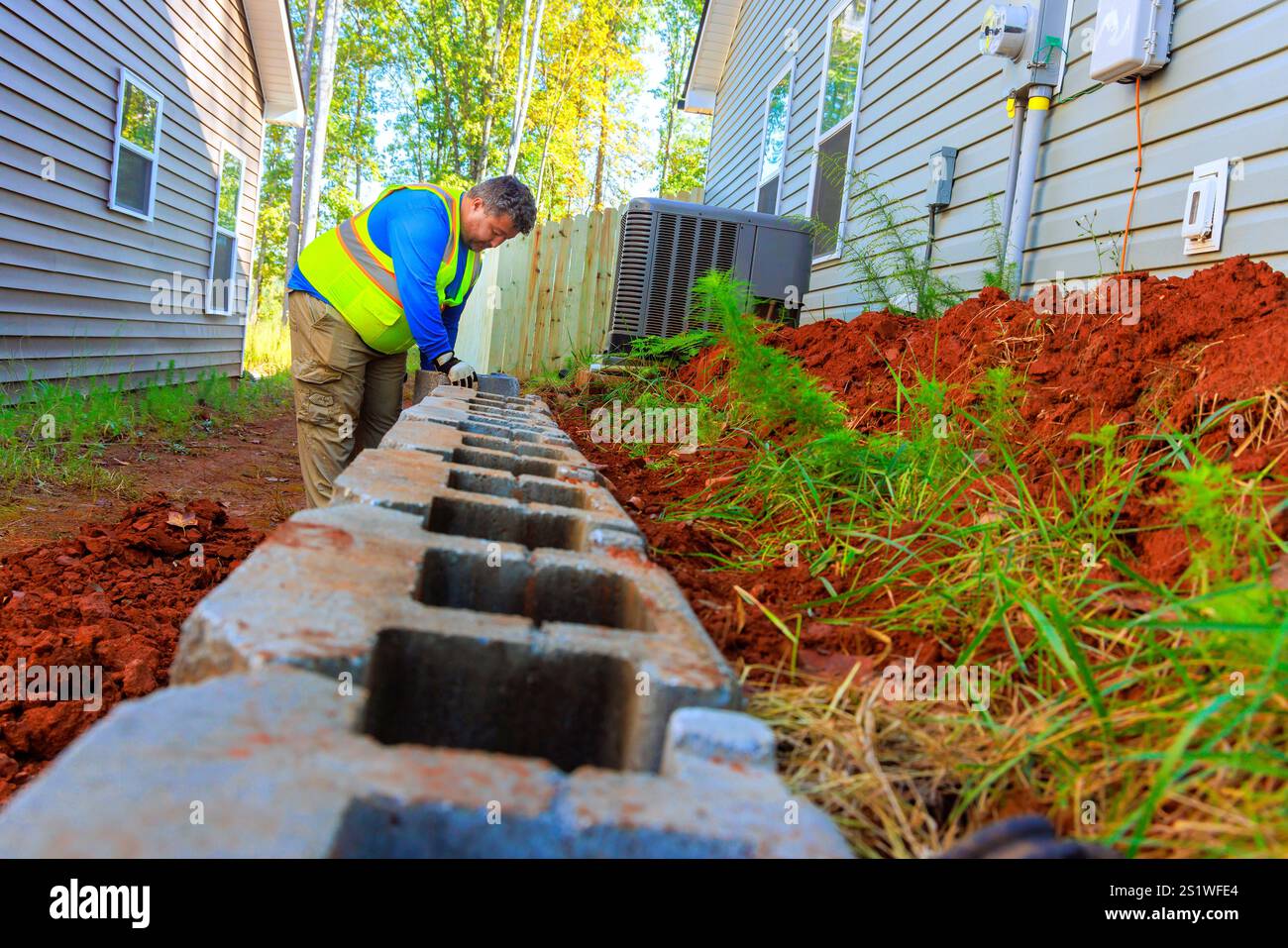 Il lavoratore si concentra sul posizionamento di blocchi a muro accanto alla casa in un tranquillo sobborgo con vegetazione lussureggiante. Foto Stock